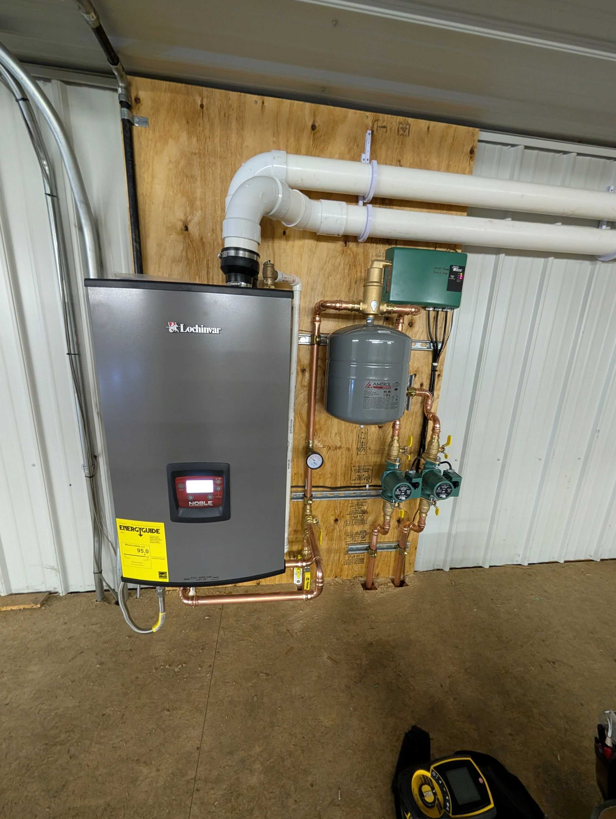 Heating system with white pipes, a green control box, a black and gray water heater, copper pipes, gauges, and a black and yellow testing device in a workshop or utility room with beige paneling and a brown carpeted floor.