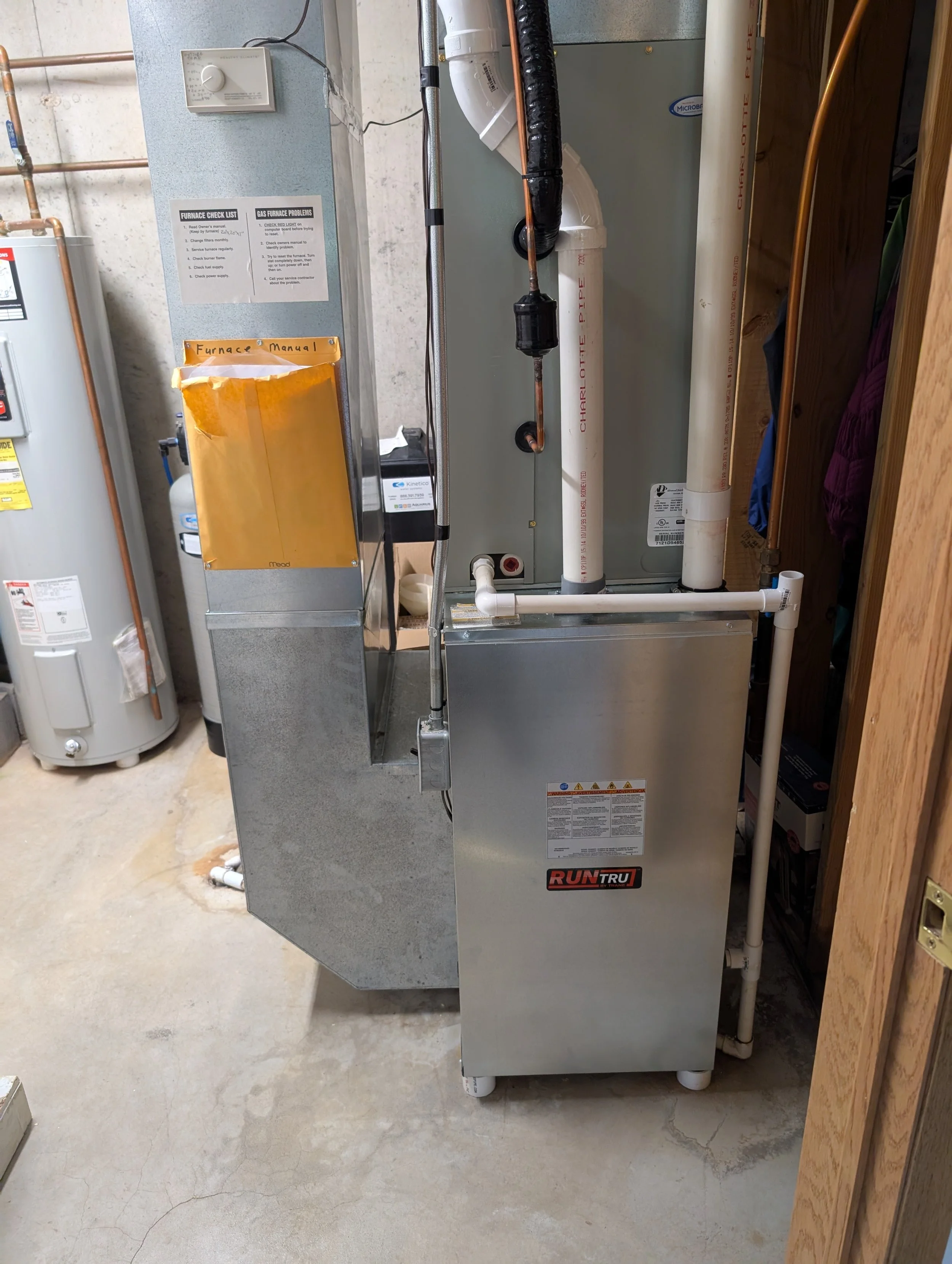 A large industrial furnace with pipes and electrical connections, situated in a basement or utility room with a concrete floor and exposed wiring.