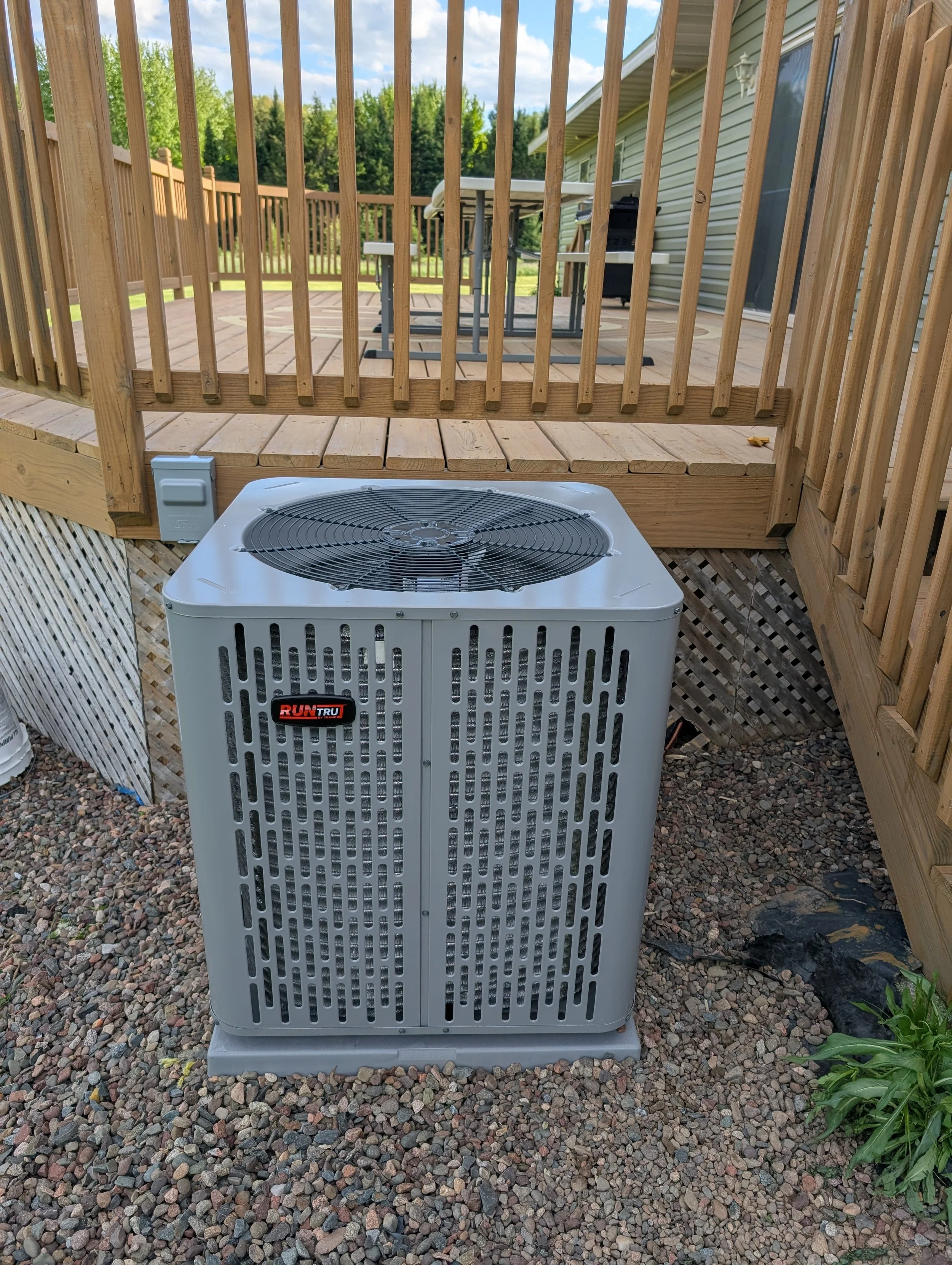 A wooden deck with a railing. An air conditioning unit is visible in the foreground. In the background, a patio table with chairs, a grill, and a house with beige siding and a sliding door. The area is outdoors with trees and a partly cloudy sky.