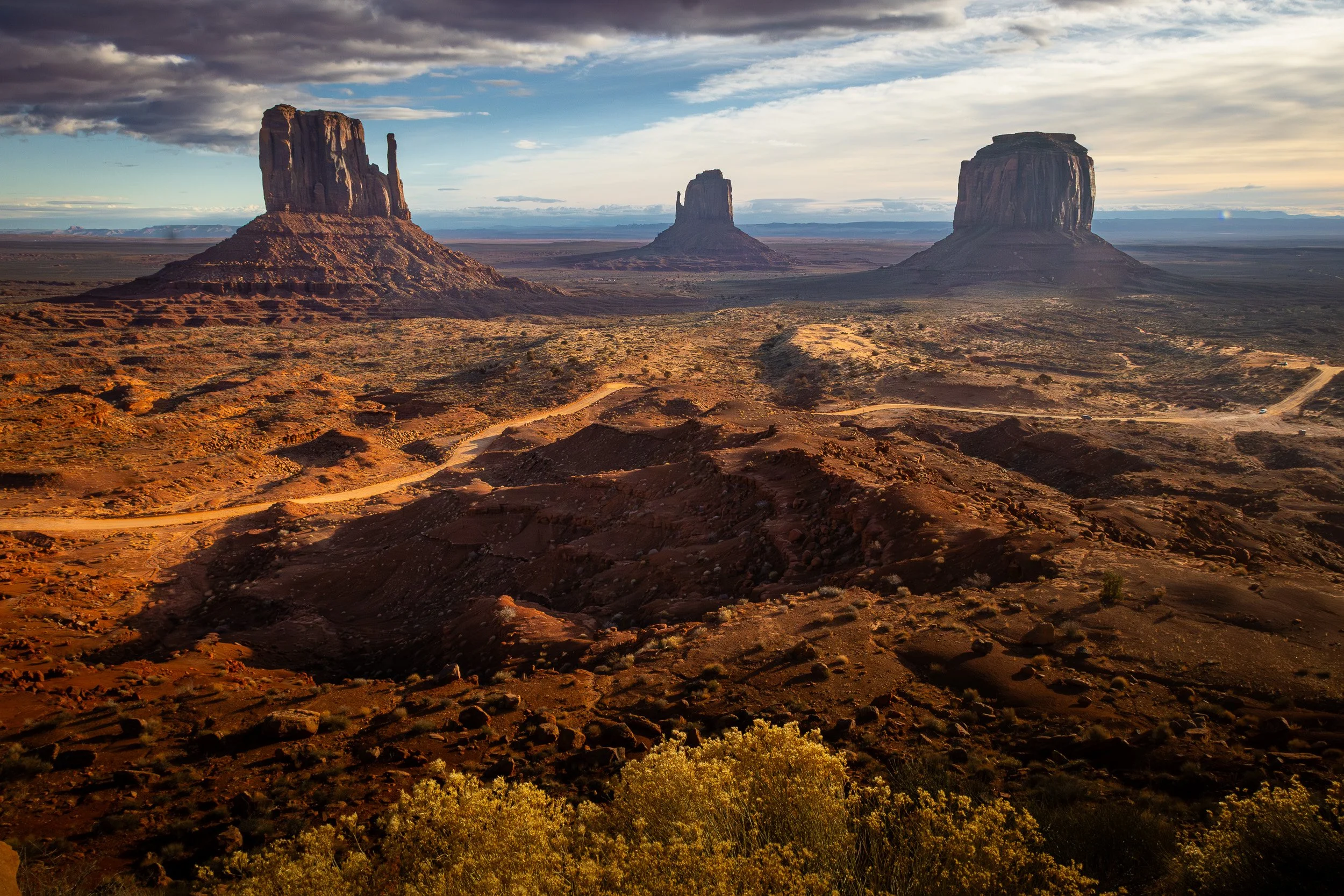 Three large mesa rock formations in a desert landscape with cloudy sky, dirt roads, and sparse vegetation.