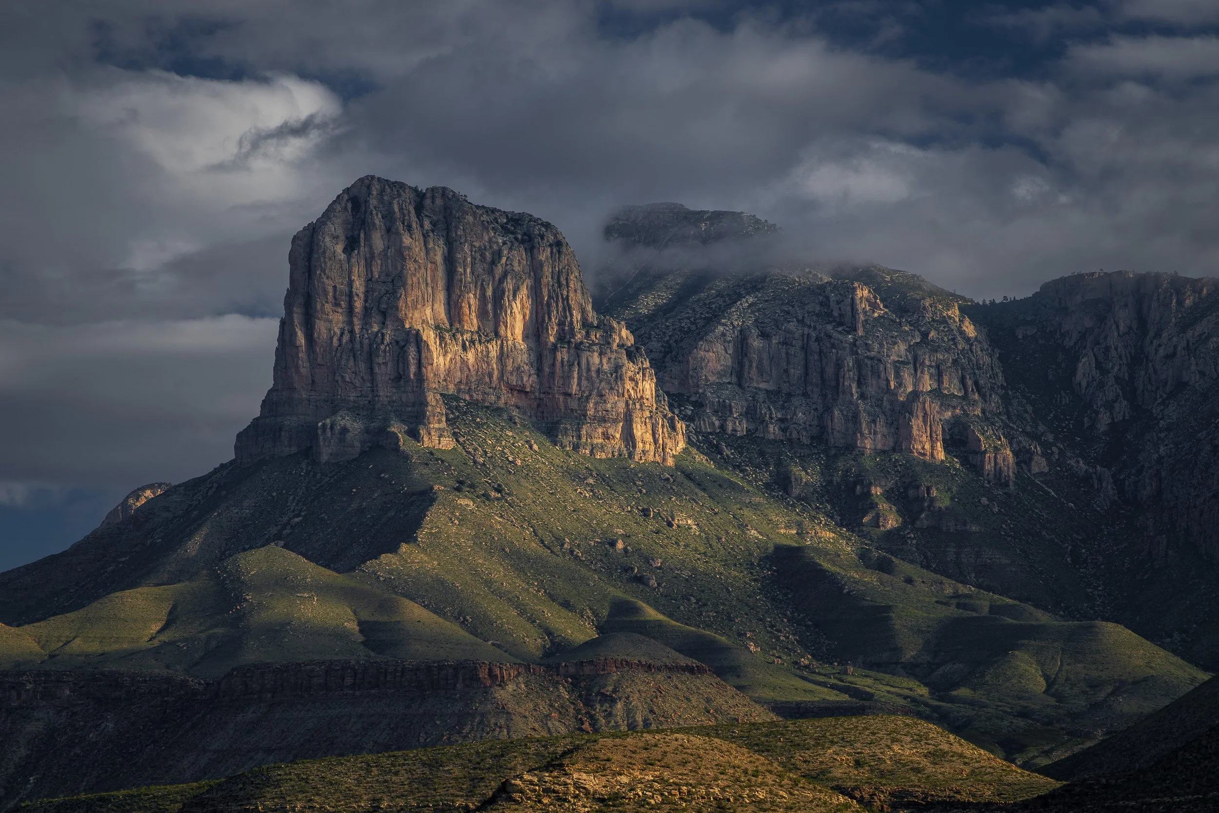 Majestic rocky mountain with green slopes at sunset, covered in clouds and mist.