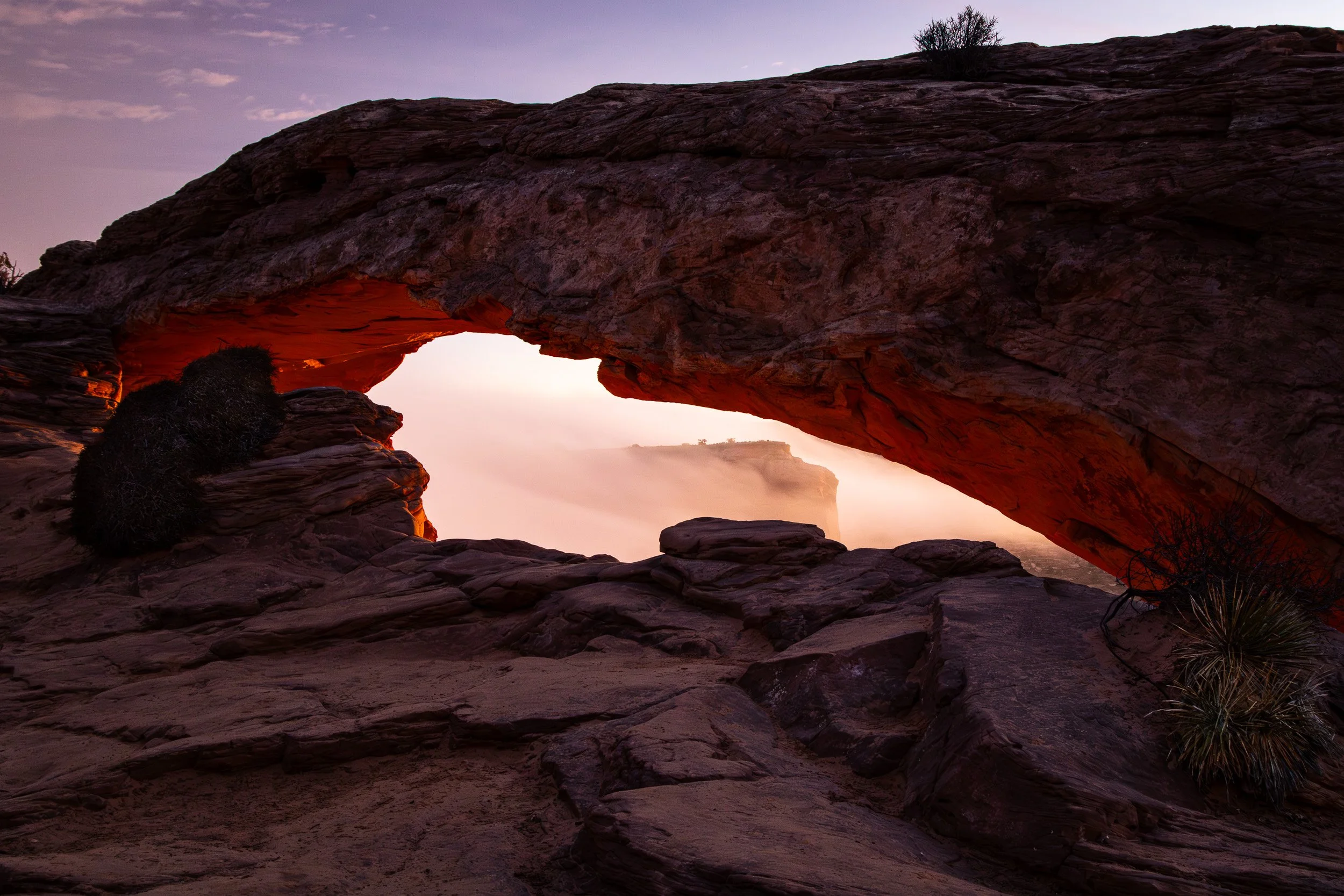 View of a natural rock formation with a large arch at sunset, illuminated by warm reddish-orange light from below, with distant fog and a purple sky.