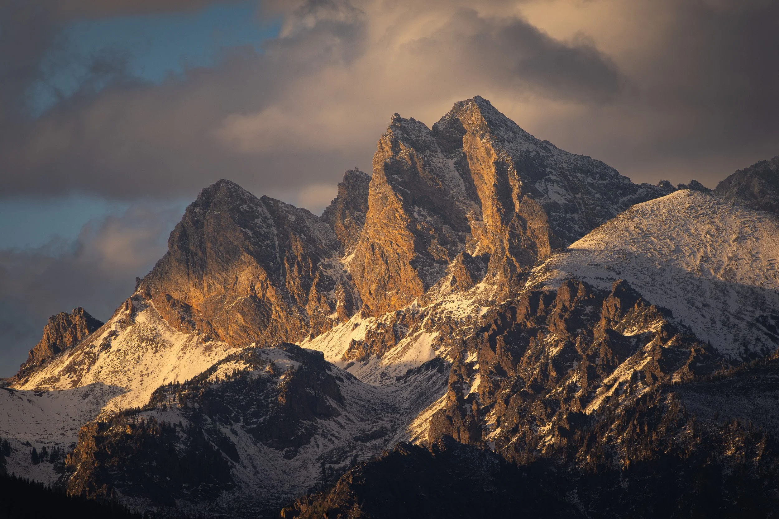 Sunlight illuminating a rugged mountain peak with snow-covered slopes and dark clouds in the sky.