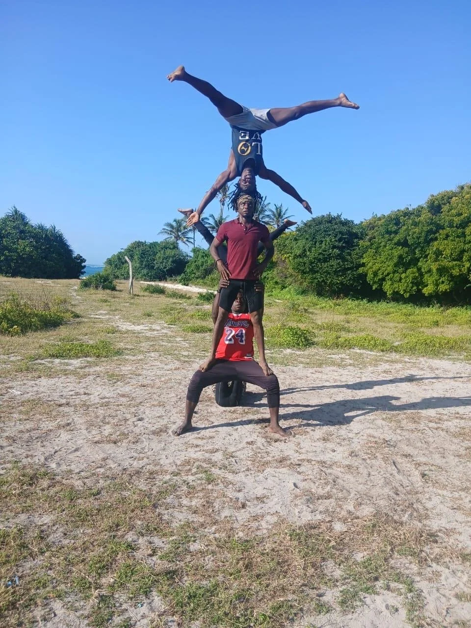 A group of four people performing a human pyramid outdoors on a dirt path with grass and trees in the background.