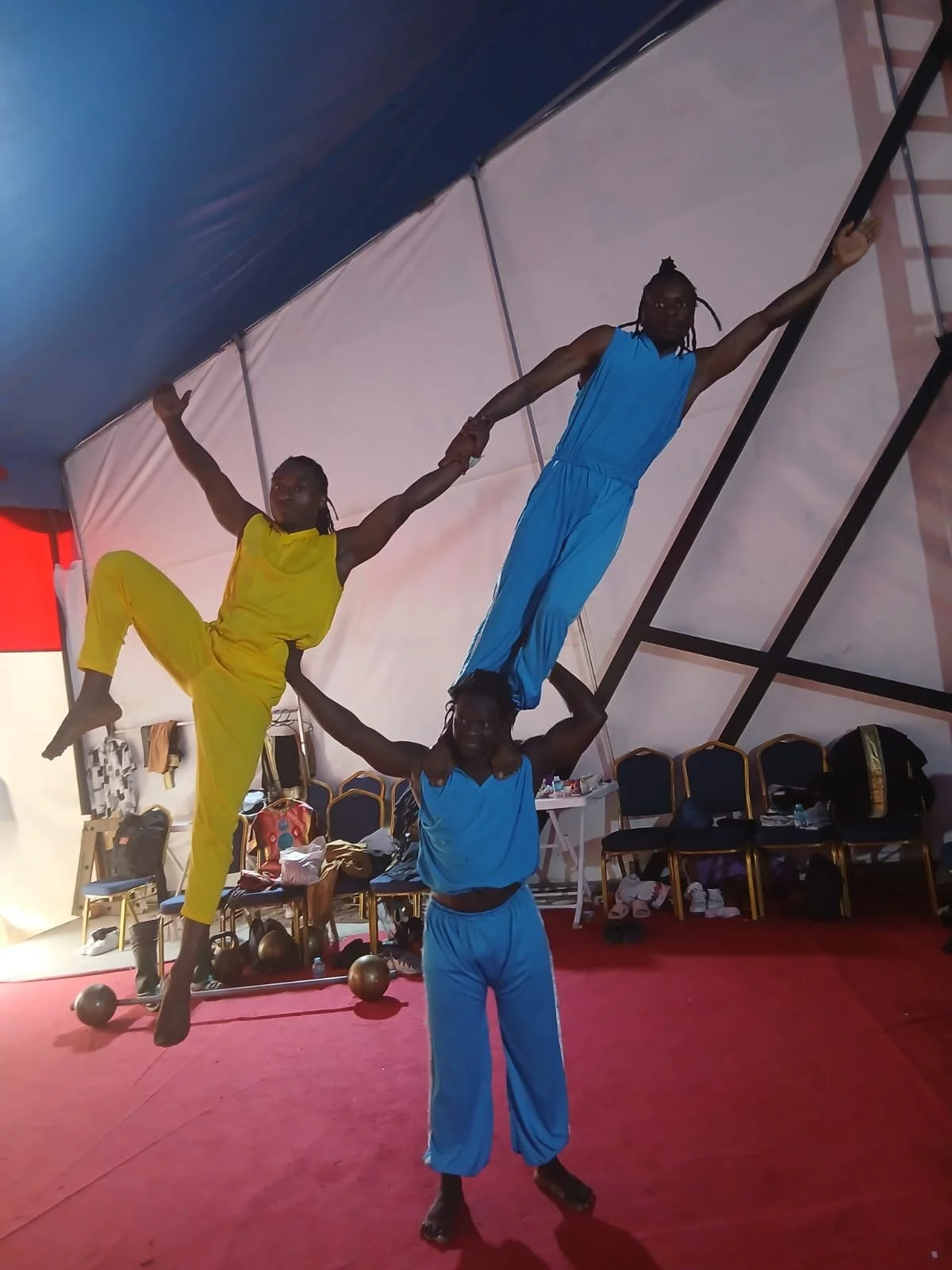 Three performers in colorful outfits executing an acrobatic pose inside a tent-like structure. One person supports another upside down while a third balances on their shoulders.