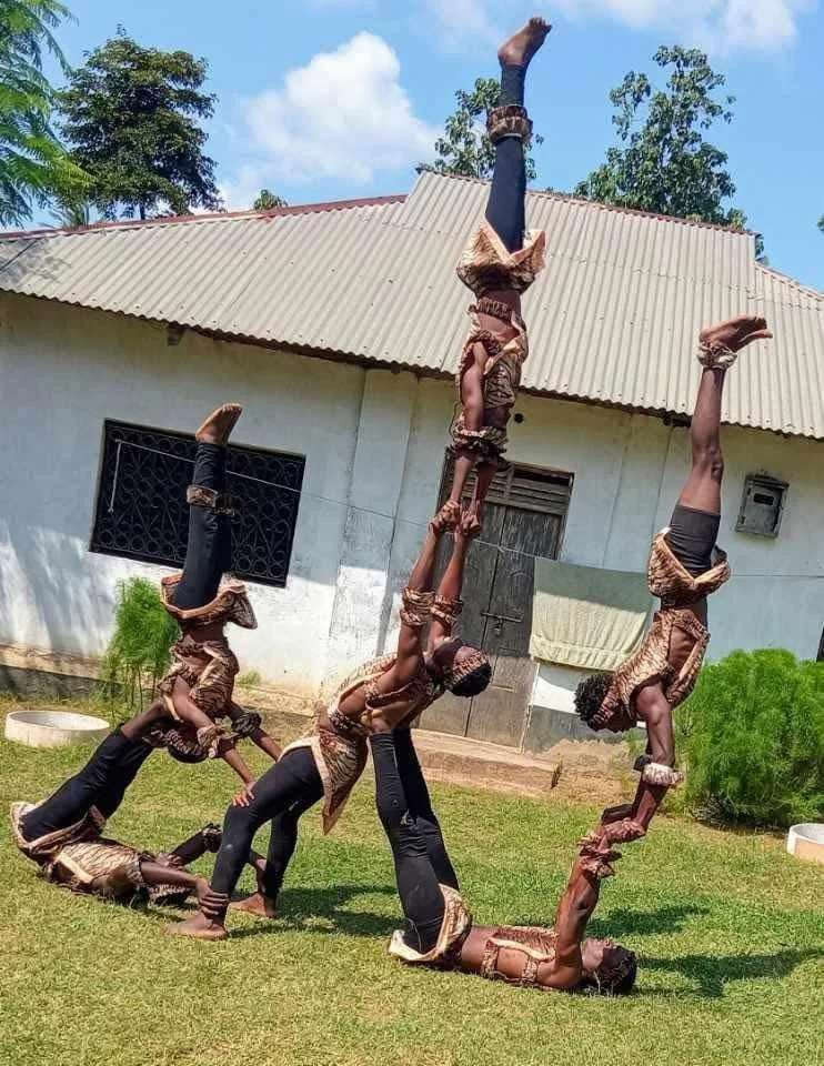 A group of children performing a coordinated acrobatic stunt outdoors, with some balancing on others' backs and arms, forming a human pyramid and line on a grassy lawn in front of a house with a corrugated metal roof.