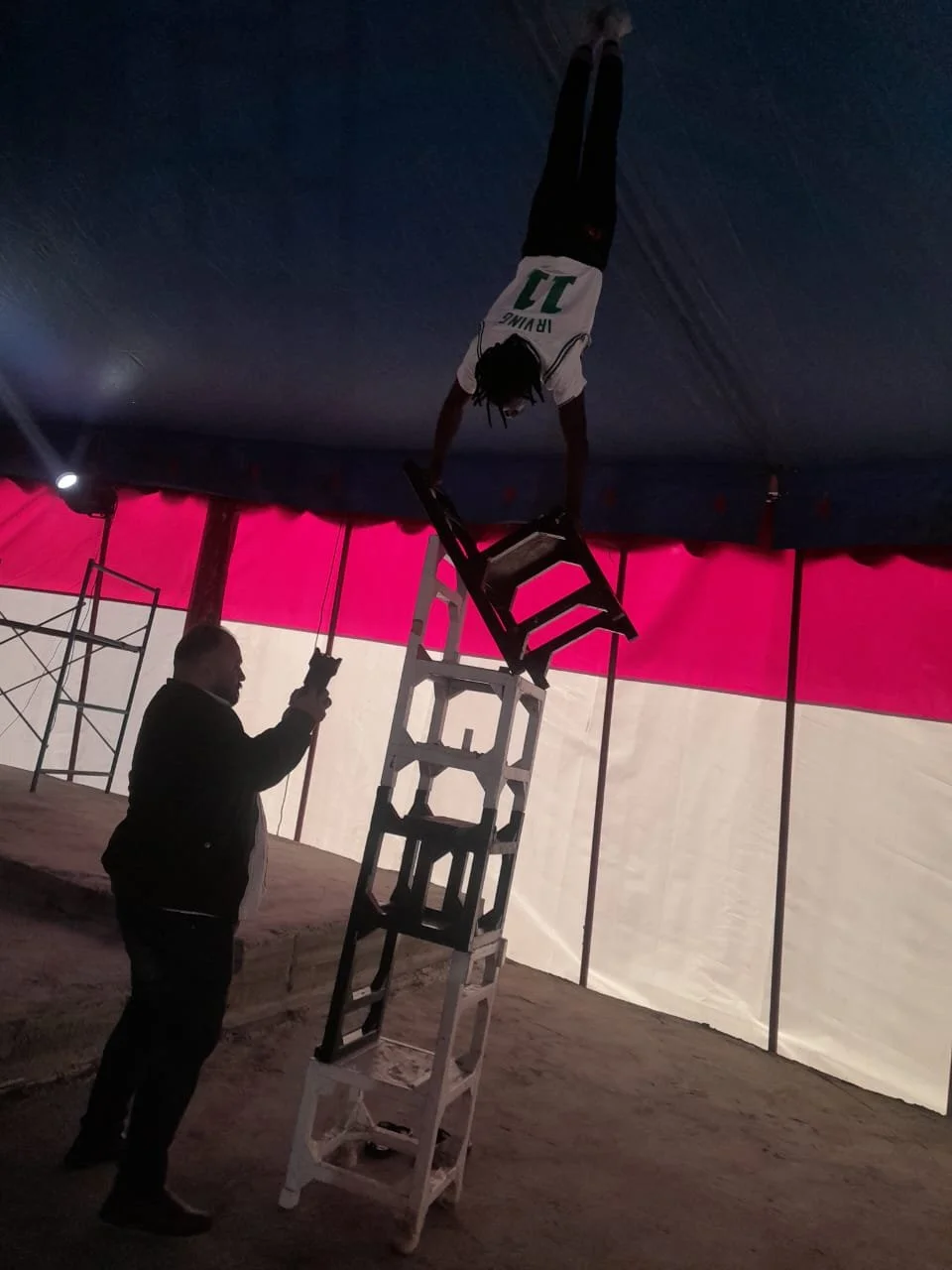 A person with dreadlocks, wearing a sports jersey with the number 11, balances on upside-down chairs stacked on a tall ladder inside a tent, while another person looks on.