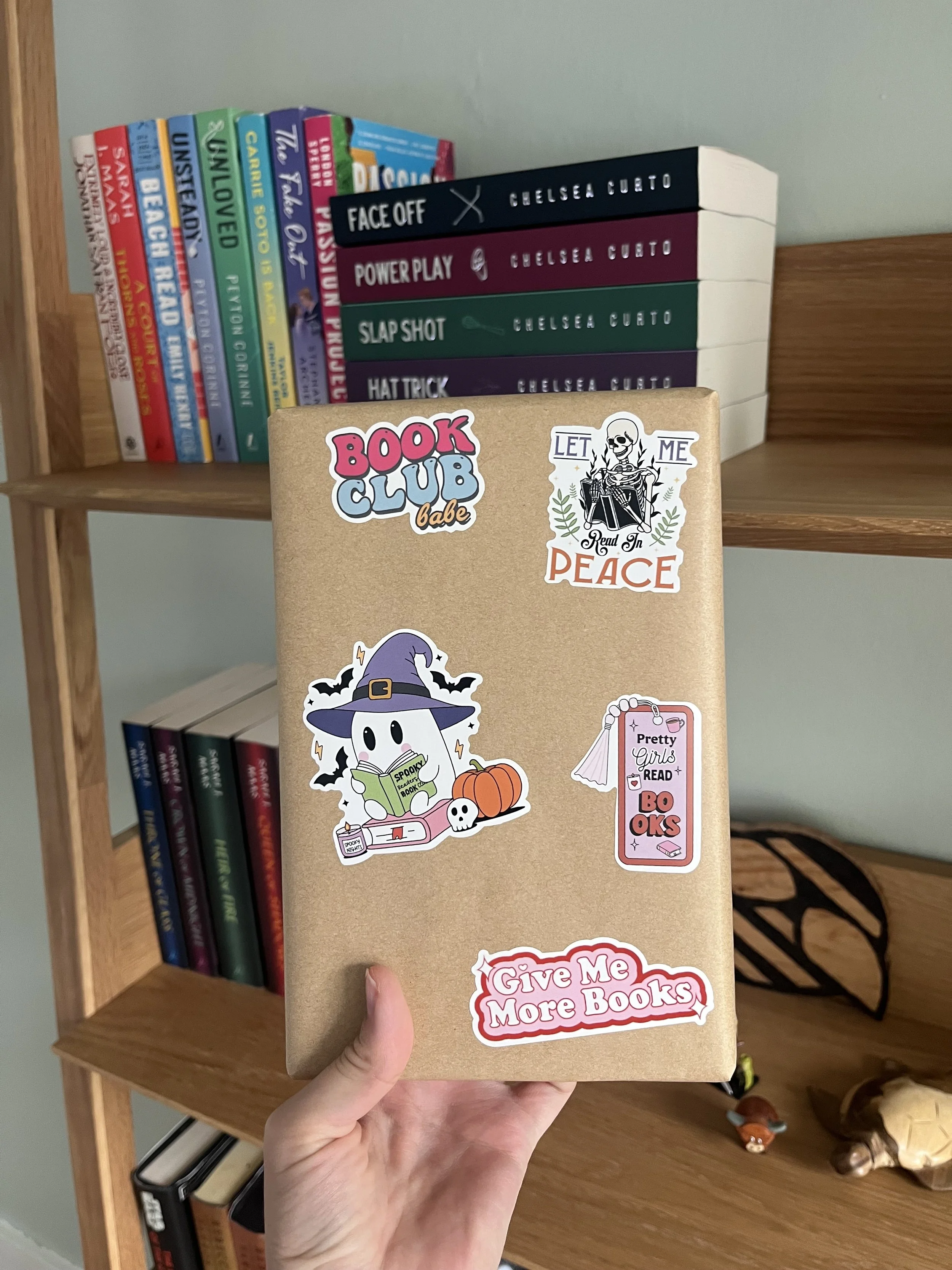 Hand holding a kraft-colored book club journal covered with book-themed stickers in front of a bookshelf.