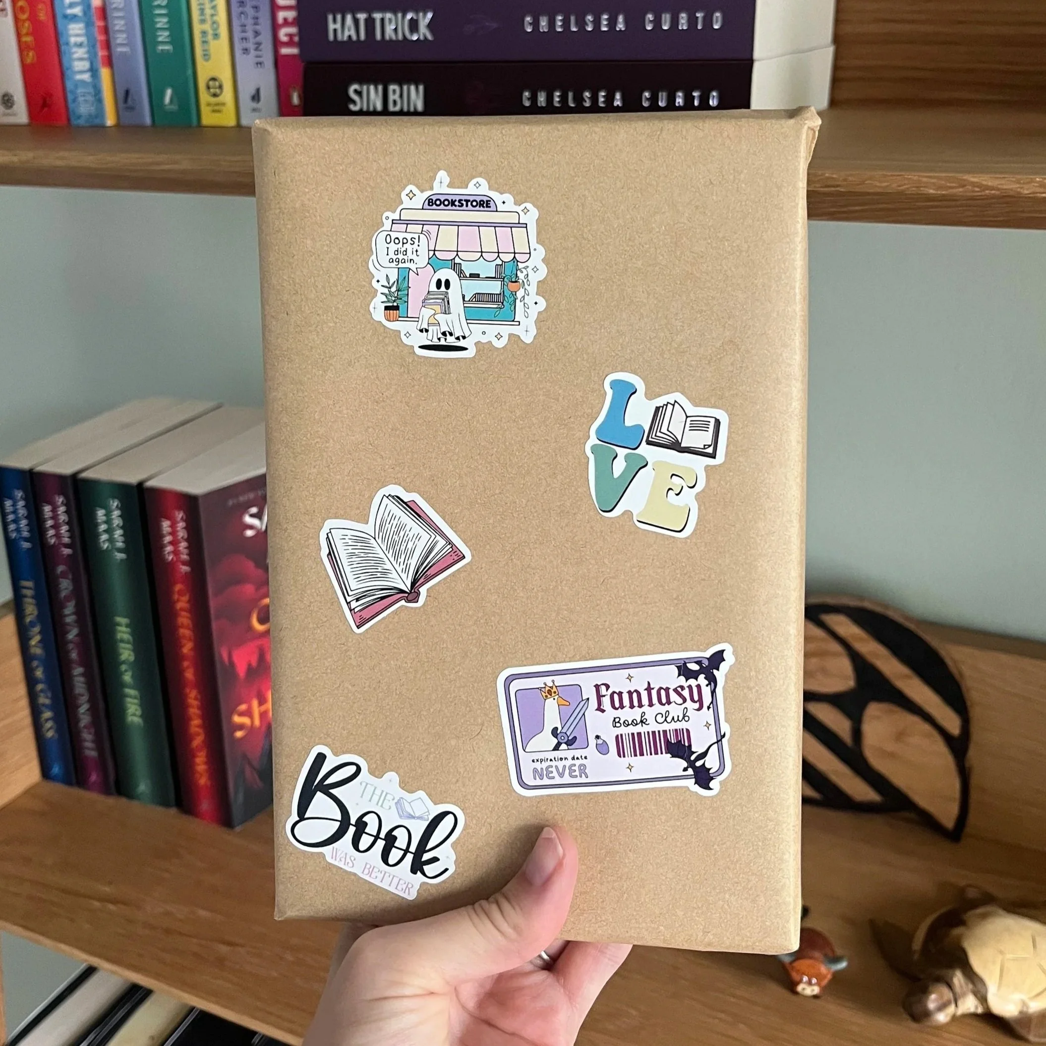 Hand holding a brown book covered with colorful book-themed stickers in front of a bookshelf.