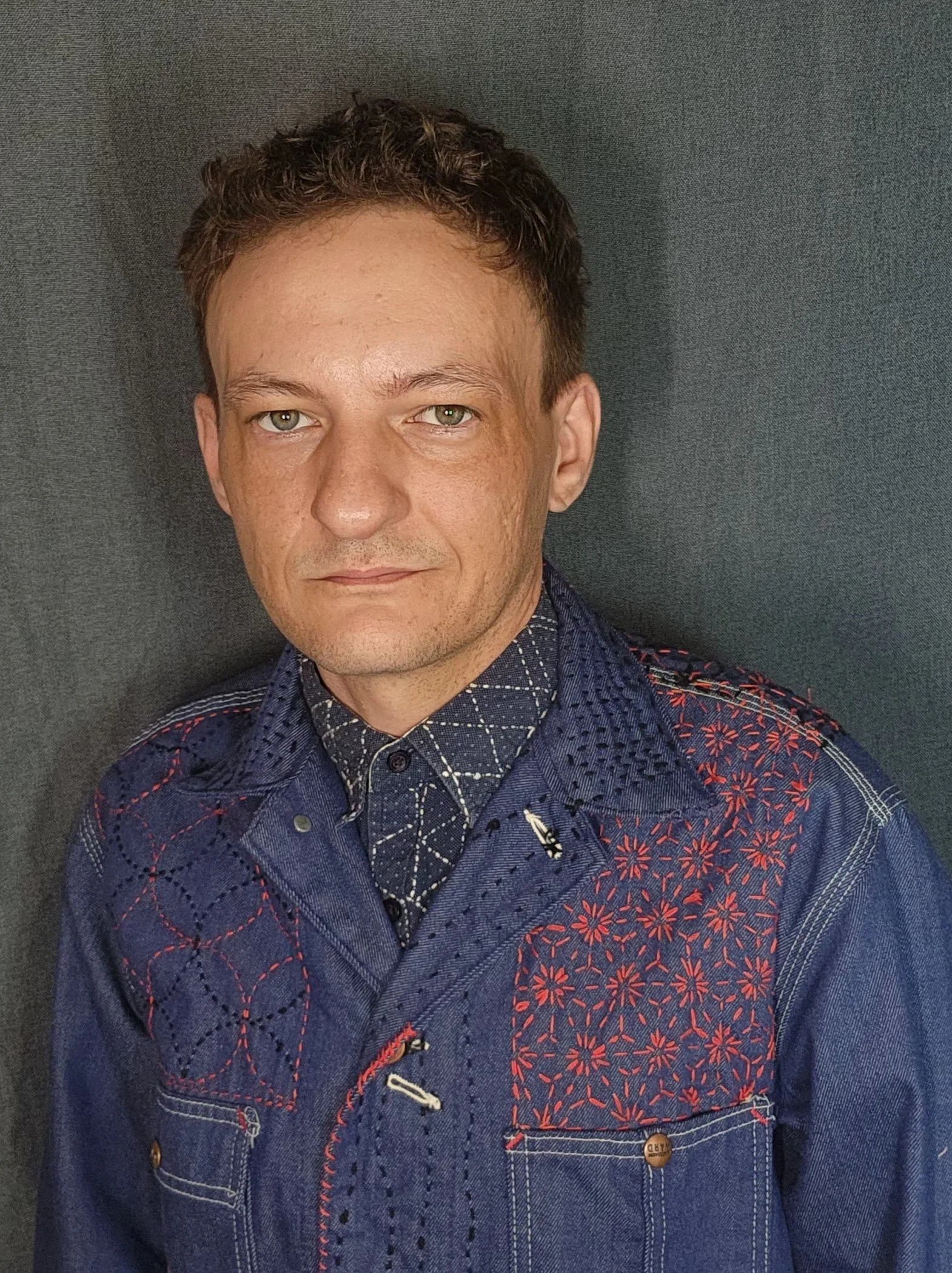 Close-up of a man with curly brown hair, wearing a blue denim jacket with intricate red embroidery, against a dark gray background.