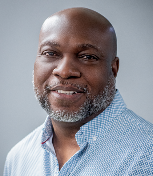 Close-up portrait of a smiling African American man with a shaved head and a graying beard, wearing a light blue collared shirt against a neutral background.