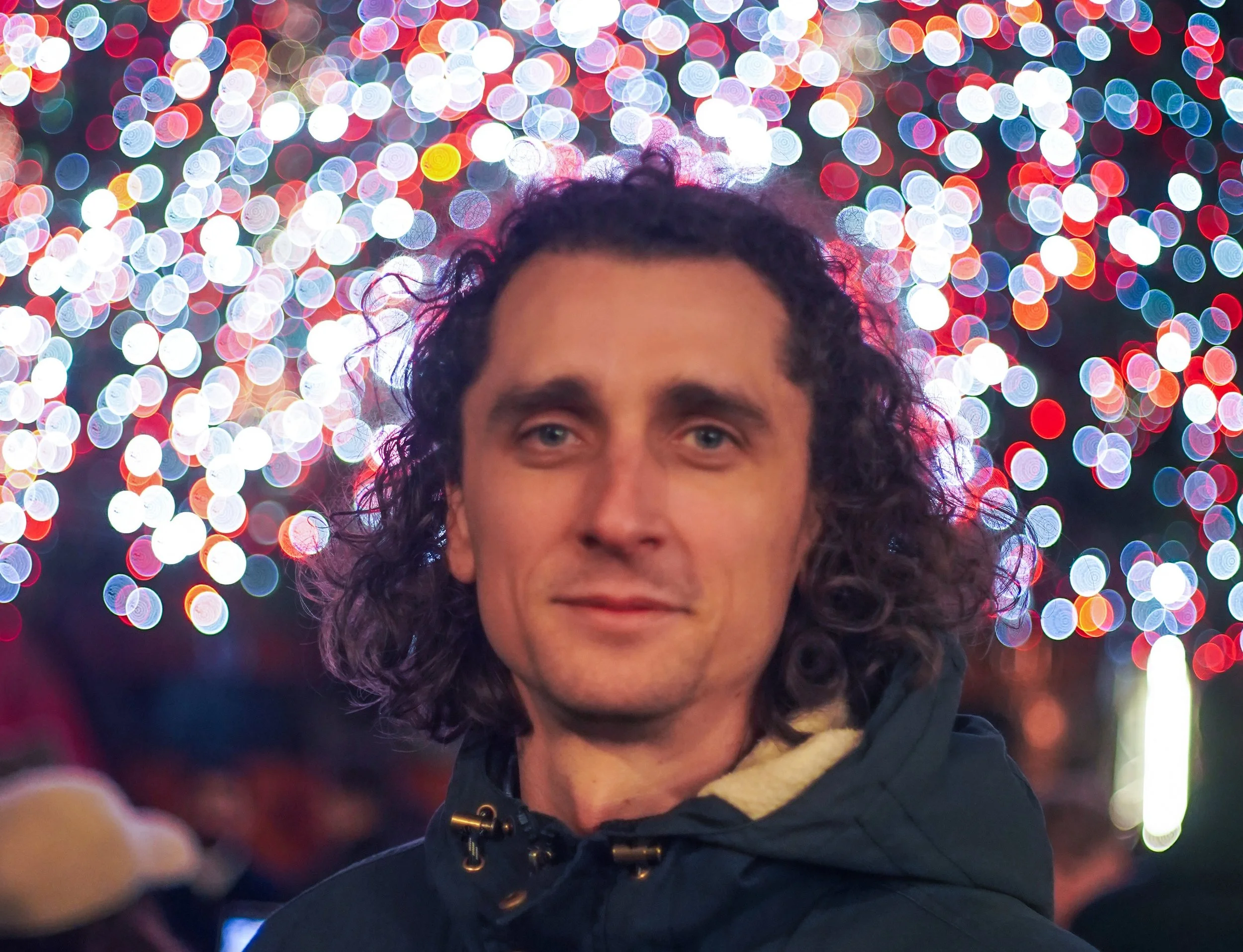 Portrait d'une personne avec des cheveux bouclés foncés, portant une veste noire, devant un fond de lumières colorées floues