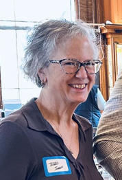 A smiling elderly woman with glasses and short gray hair, wearing a dark top and a name tag, sitting indoors with a window and wooden furniture in the background.