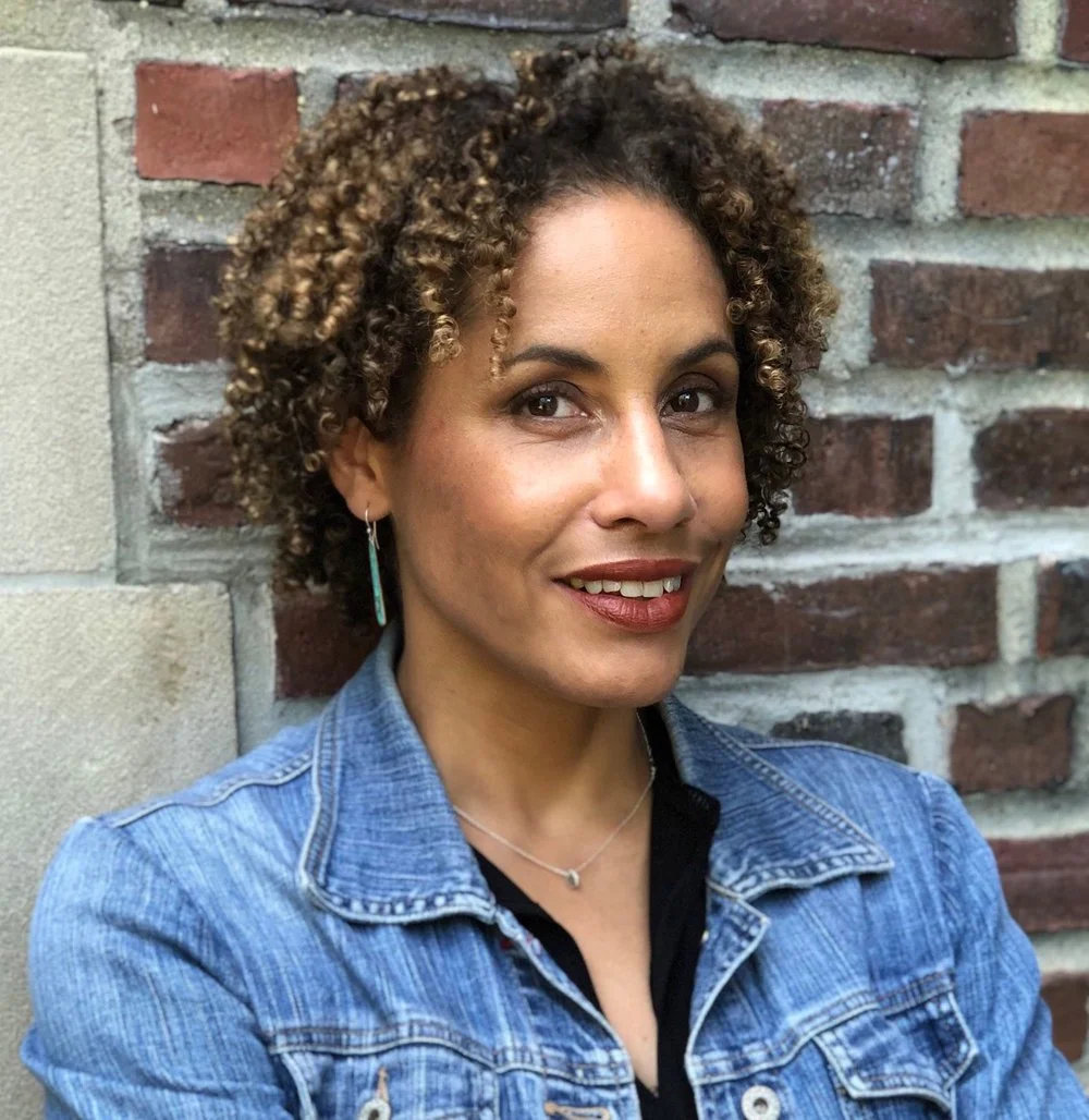 A woman with curly hair wearing a denim jacket, silver jewelry, and smiling, standing against a brick wall.