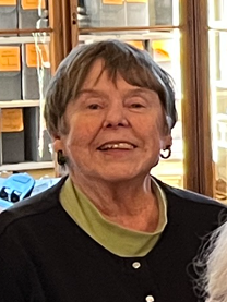 Smiling elderly woman with short brown hair, wearing a black top and a green shirt underneath, standing indoors near windows.