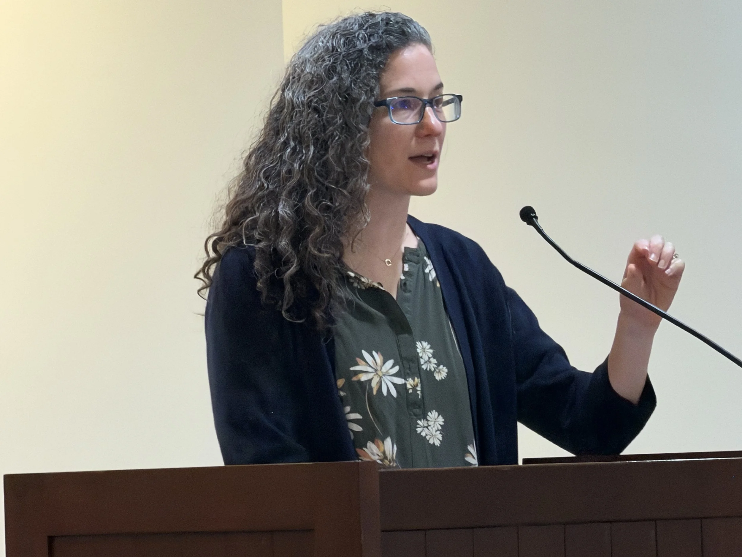 A woman with curly gray hair and glasses speaking at a podium with a microphone.