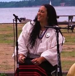 Woman in a white graduation gown sitting outdoors near a lake, smiling.