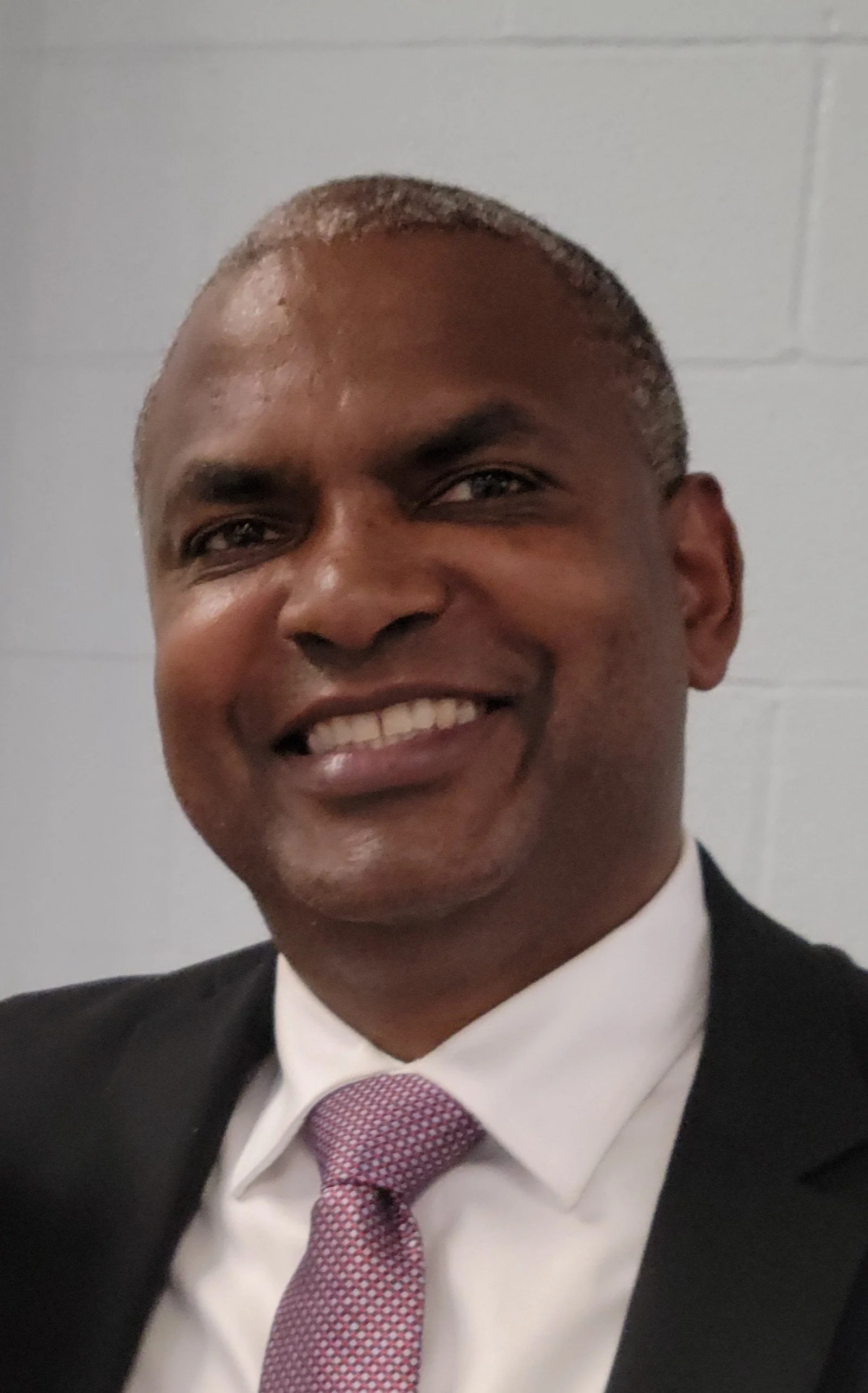 Smiling man in a black suit with a pink tie, white shirt, and gray hair, posed against a plain white background.
