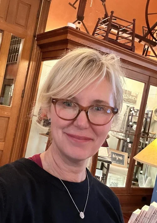 A woman with short blonde hair wearing glasses and a black top, indoors with wooden furniture and glass display cabinets in the background.
