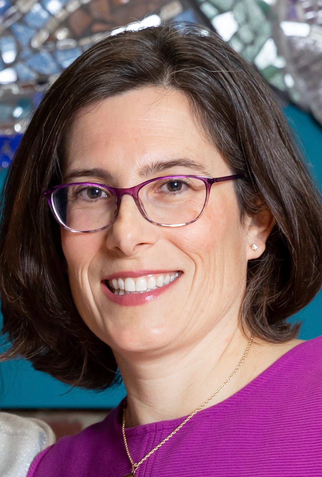 Close-up portrait of a woman with brown hair wearing purple glasses and a purple top, smiling in front of a colorful abstract background.
