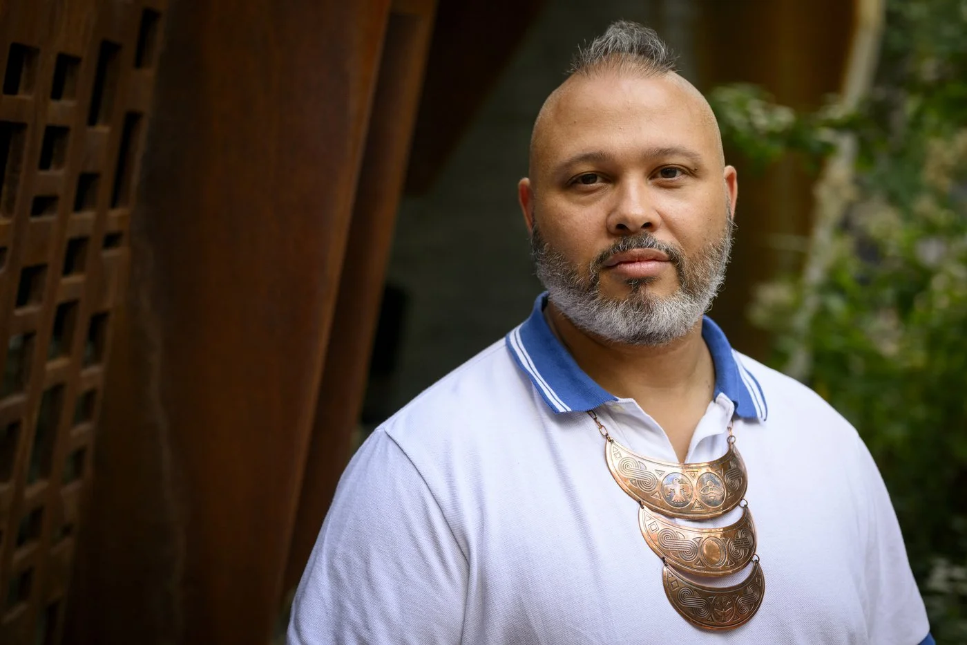 Portrait of a man with a gray beard and mohawk hairstyle wearing a white polo shirt with a blue collar and a large ornate necklace, standing outdoors near a wooden structure and greenery.