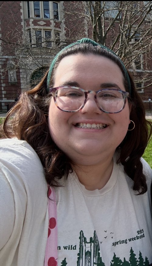 Young woman with glasses smiling outdoors in front of a brick building and leafless trees on a sunny day.