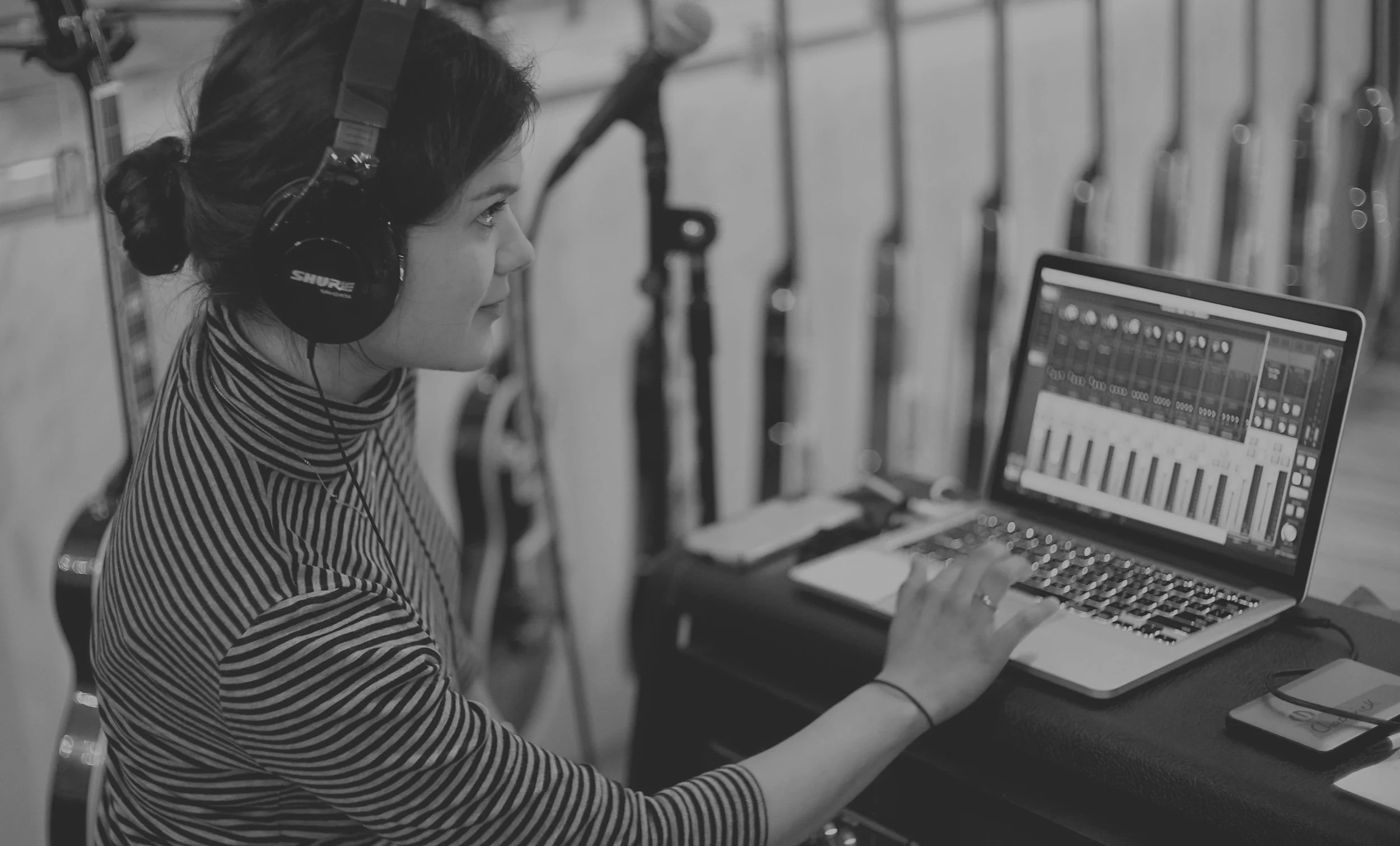 A woman wearing headphones working on a laptop with music production software in a music studio.