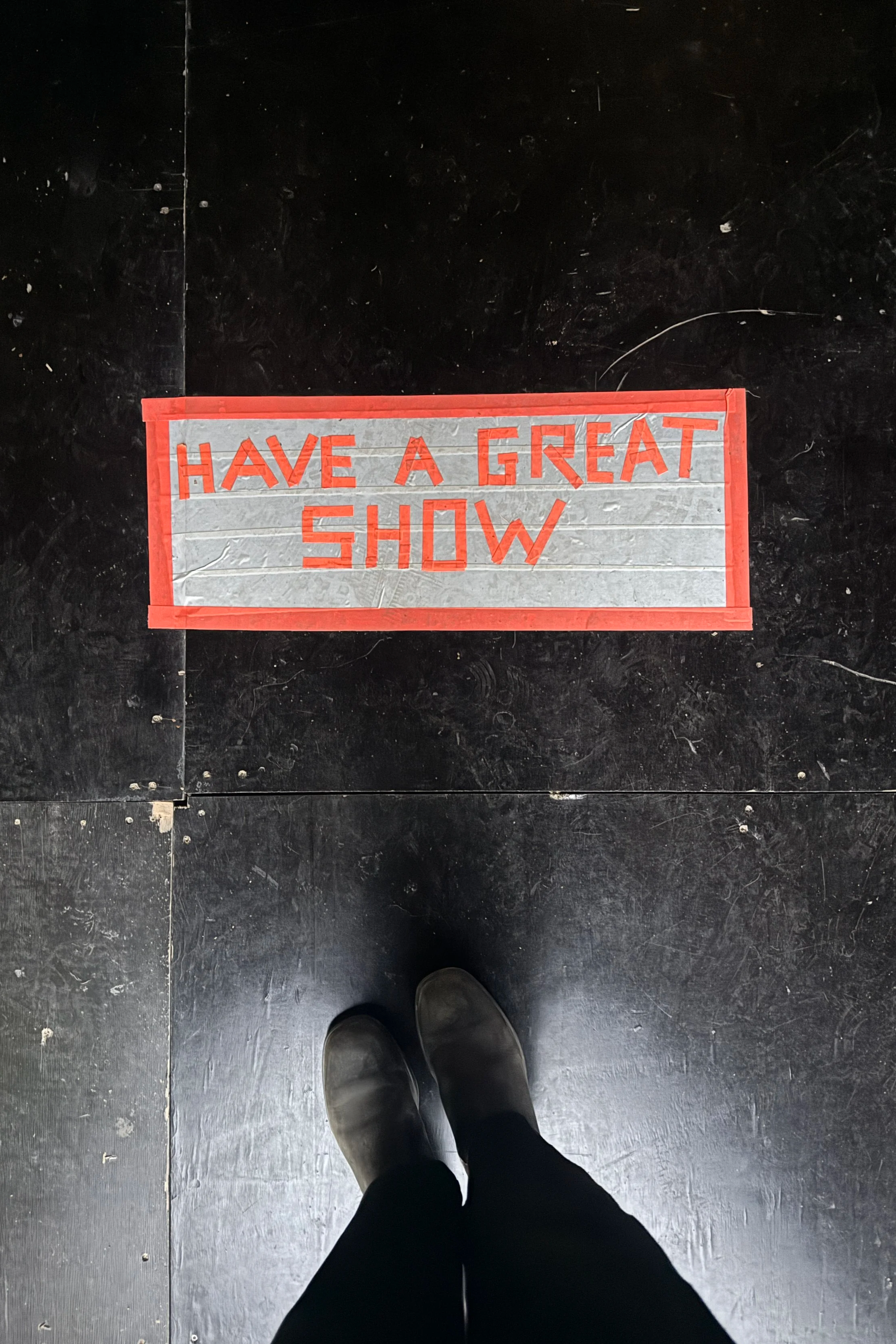 A person standing on a black tiled floor, wearing black shoes and dark pants, looking down at a rectangular sign that says 'HAVE A GREAT SHOW' in red letters on a gray background with a red border.