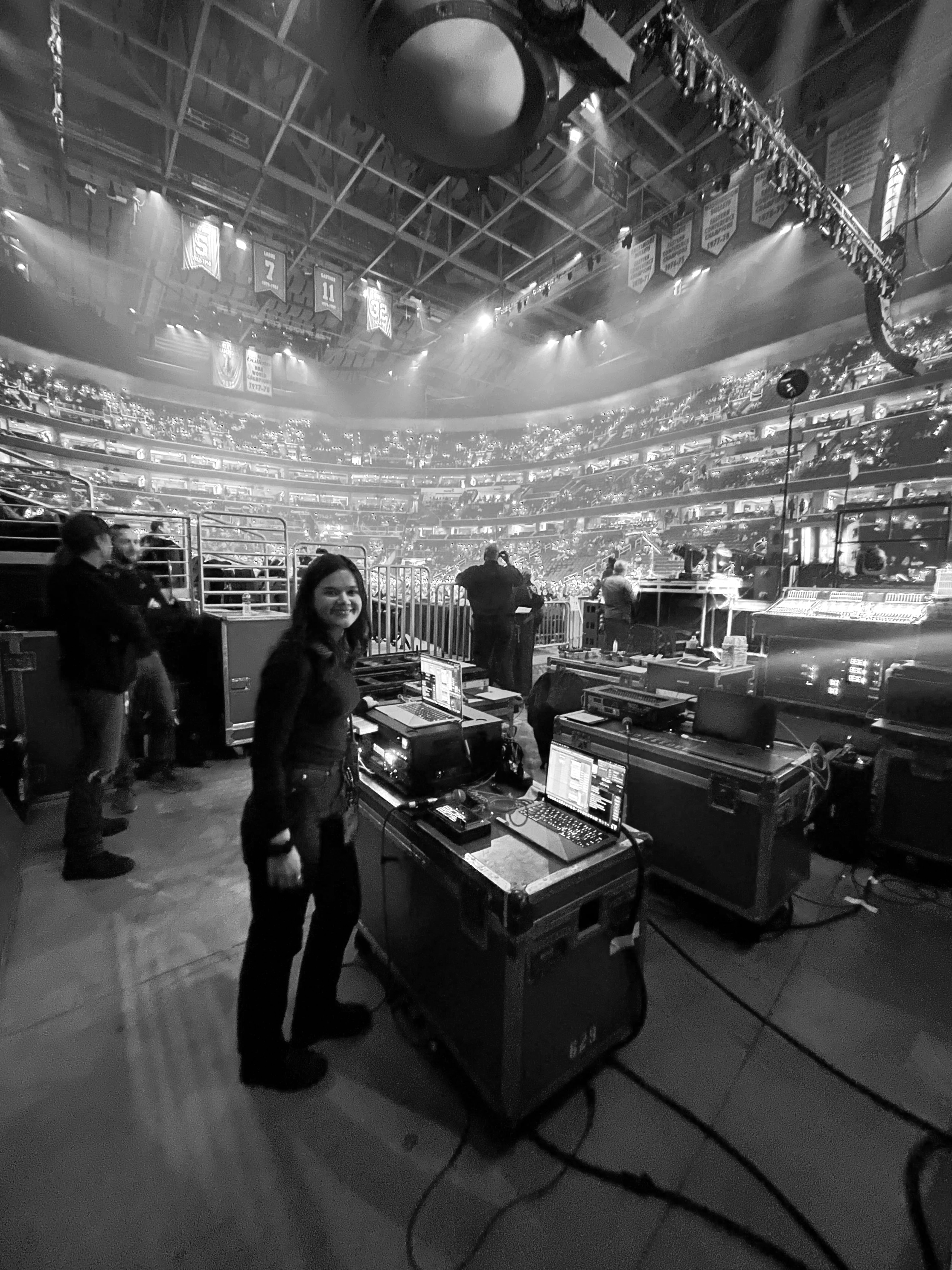 The photo shows a large indoor arena filled with spectators, with several banners hanging from the ceiling. In the foreground, there are people working at a control station with computers and audio equipment. The arena appears to be set up for a sports event or concert, with the seating area and stage visible in the background.