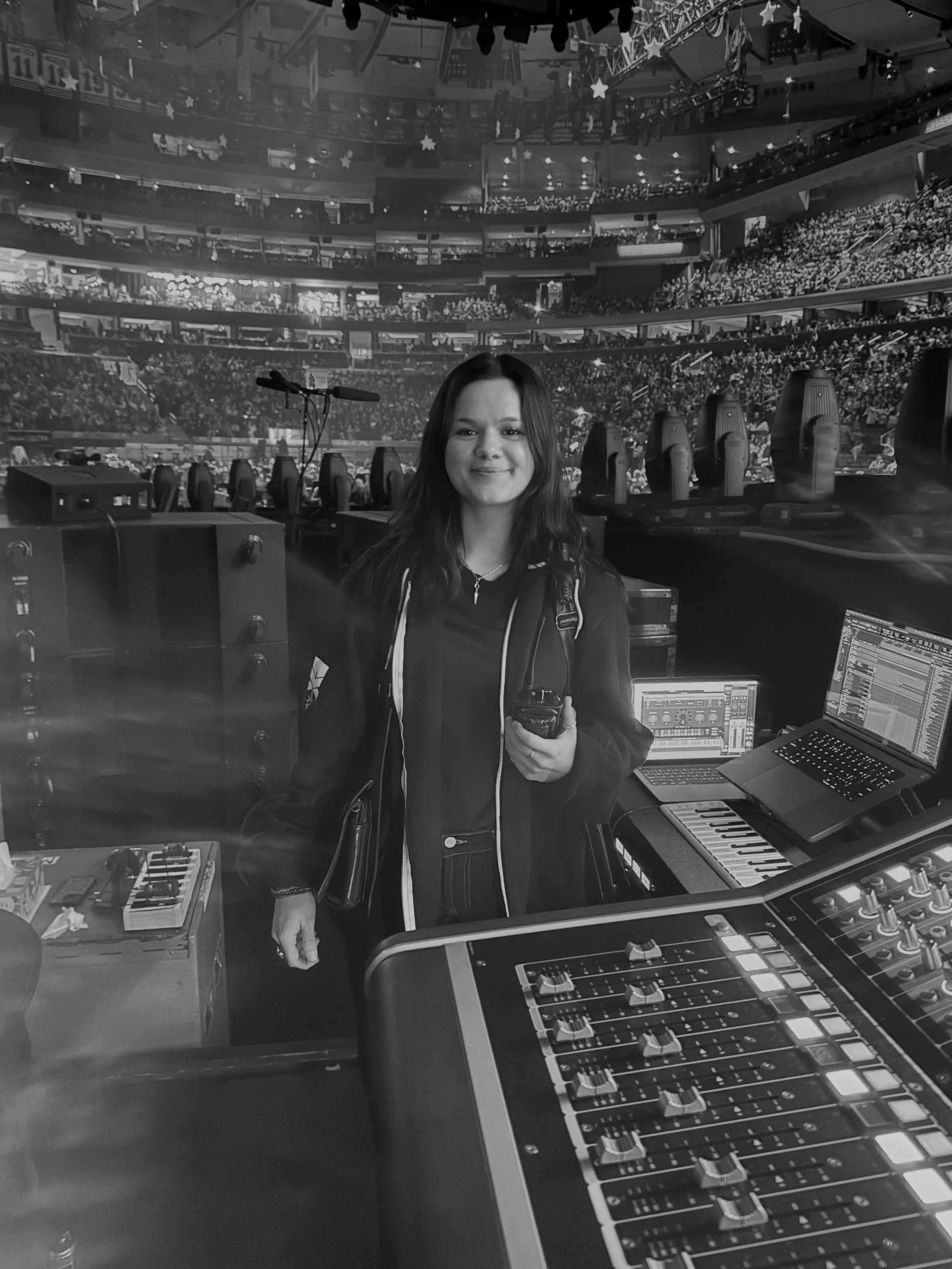 A woman standing in front of a stage with a large indoor arena filled with audience in the background. She is holding a walkie-talkie, and there is DJ or music equipment in front of her, including a mixer, laptop, keyboard, and sound control devices.