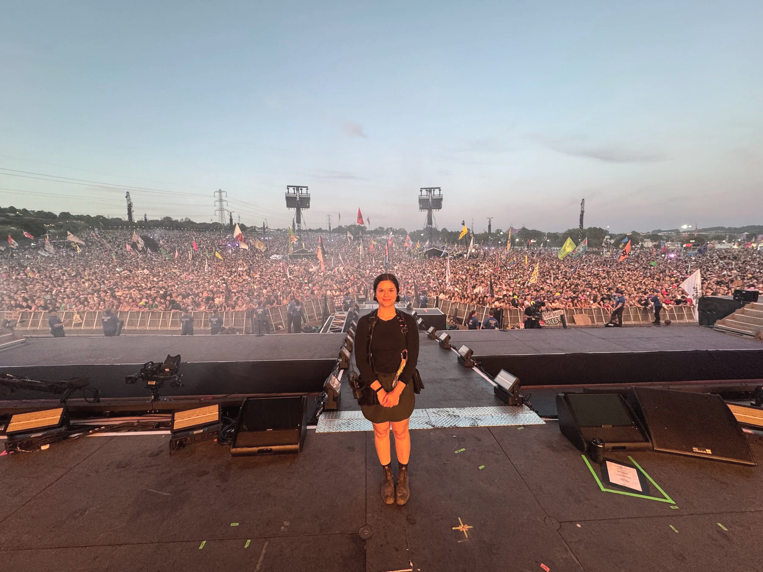 A young woman with dark hair, wearing a black long-sleeve shirt and brown boots, standing on a stage with a large crowd in the background at an outdoor concert or festival during dusk.