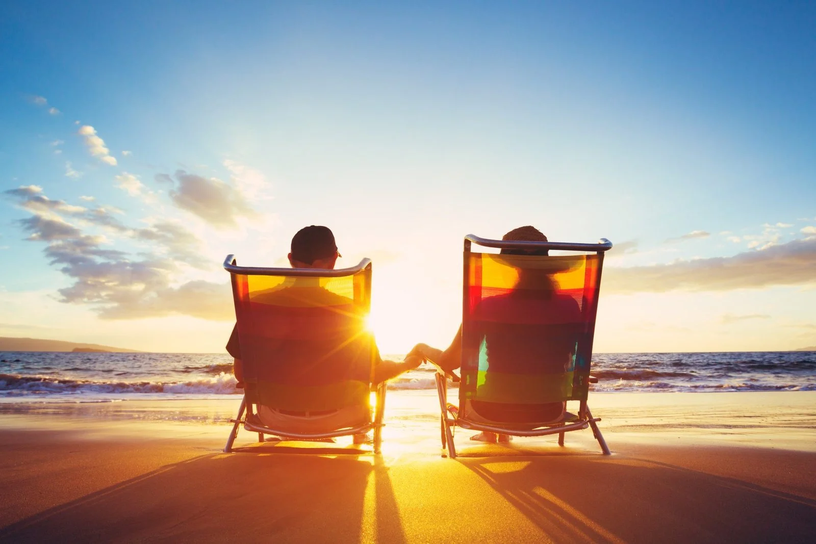 Two people sitting in beach chairs holding hands at sunset on the beach, facing the ocean with the sky and clouds in the background.