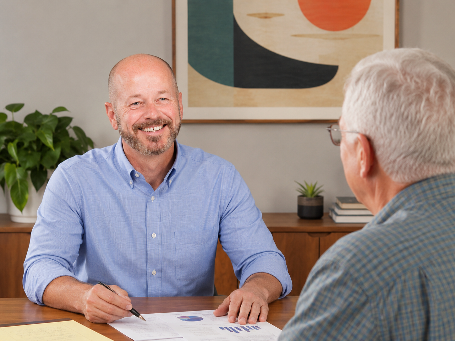 Two men having a conversation at a desk with documents, one smiling and holding a pen, the other with white hair and glasses. A plant and framed artwork are in the background.
