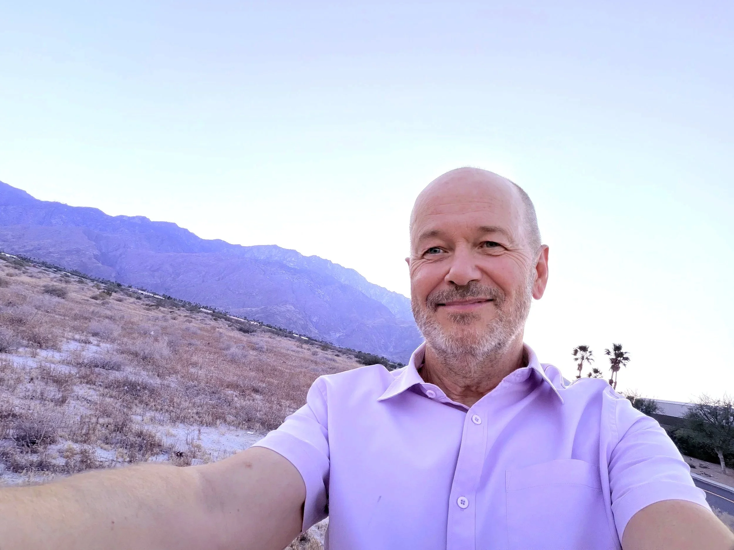 A middle-aged man with a beard and short hair, wearing a white shirt, is taking a selfie outdoors in a desert landscape with mountains in the background during the daytime.