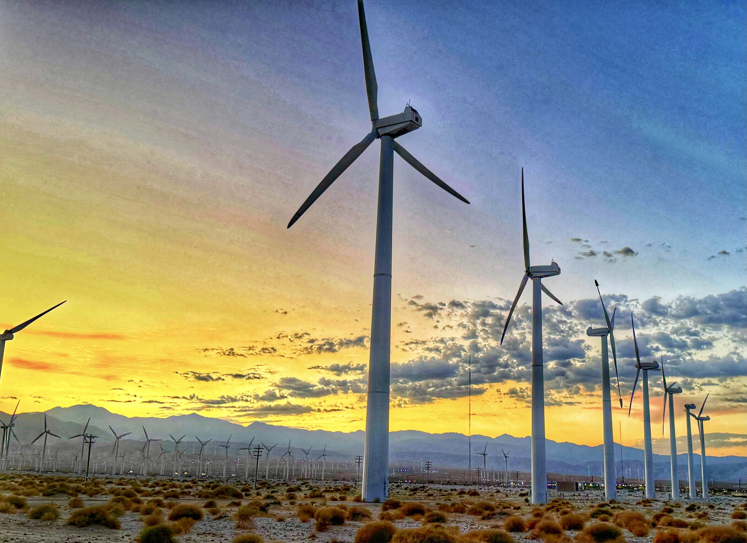 A field of wind turbines under a colorful sunset sky with mountains in the background.