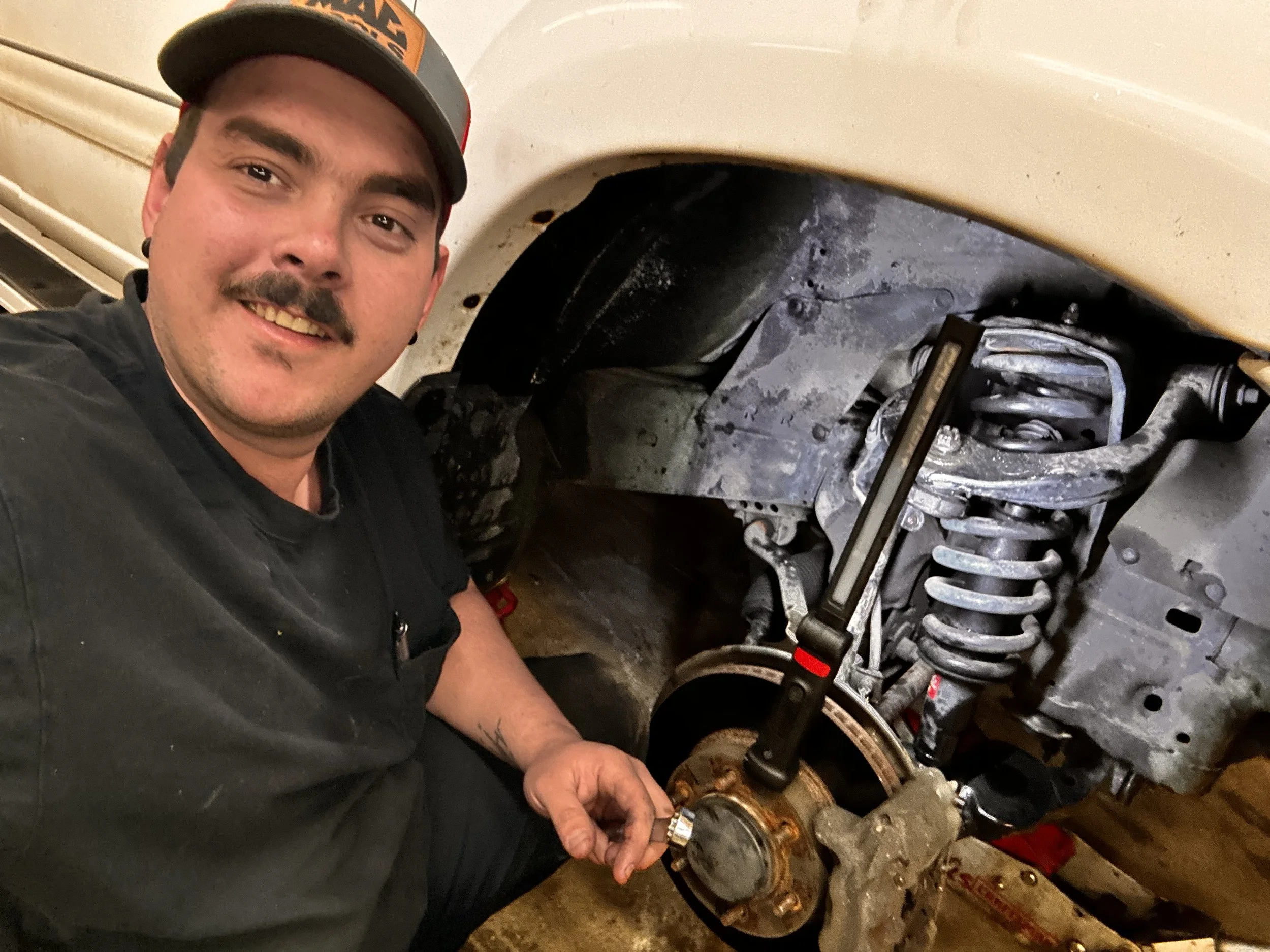 A man working on a vehicle's suspension system, holding a tool near the wheel well with exposed suspension components including a coil spring and brake rotor.