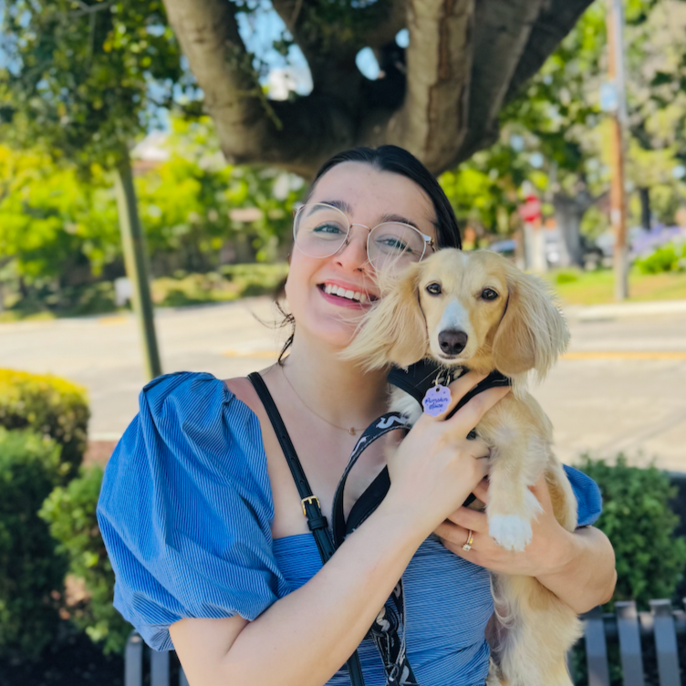 A smiling woman with glasses holding a small, light-colored dog with long ears and a collar, standing outdoors under a large tree.