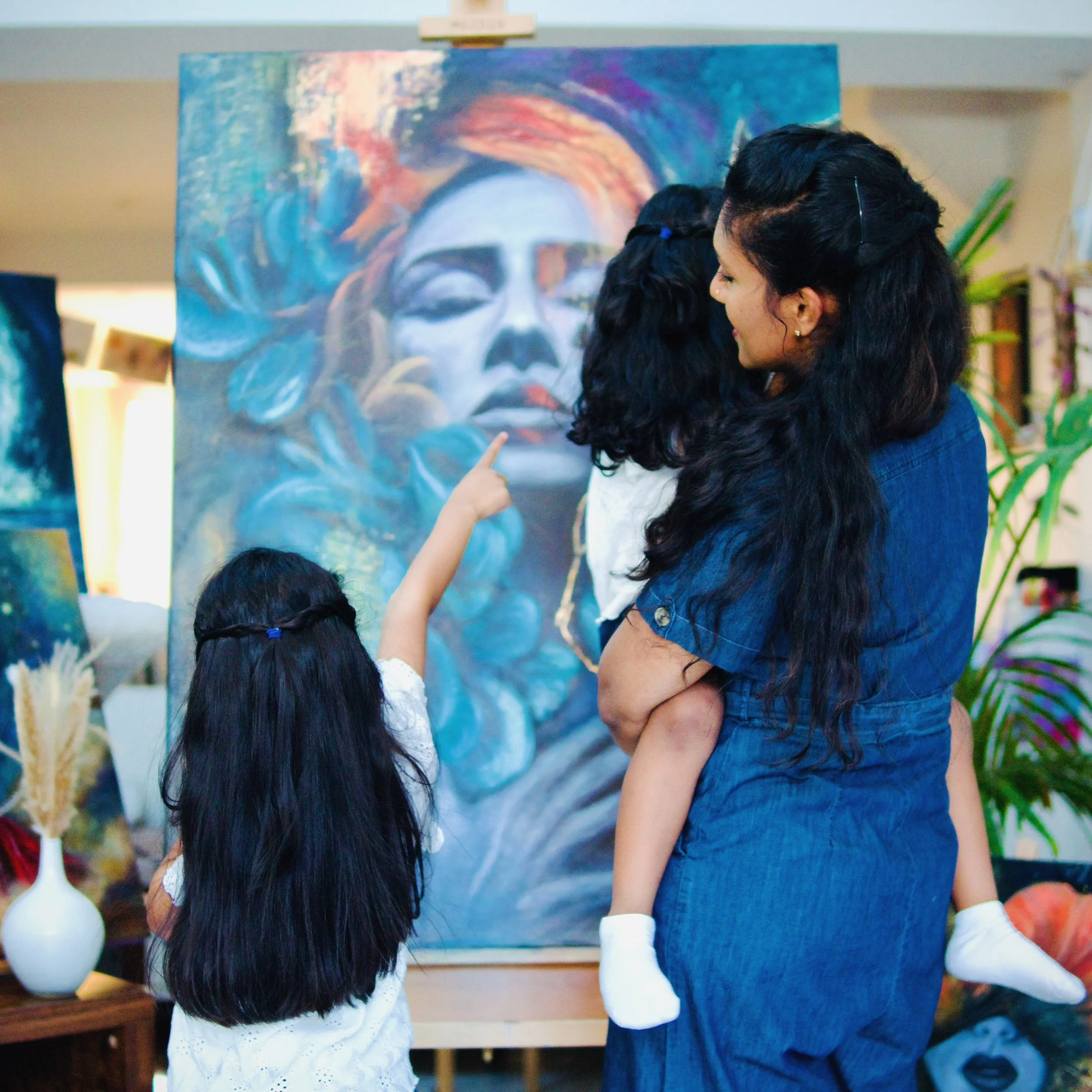 Two young girls with long dark hair, one wearing a white dotted shirt and the other a denim shirt, standing in front of a colorful abstract painting, pointing at the artwork.