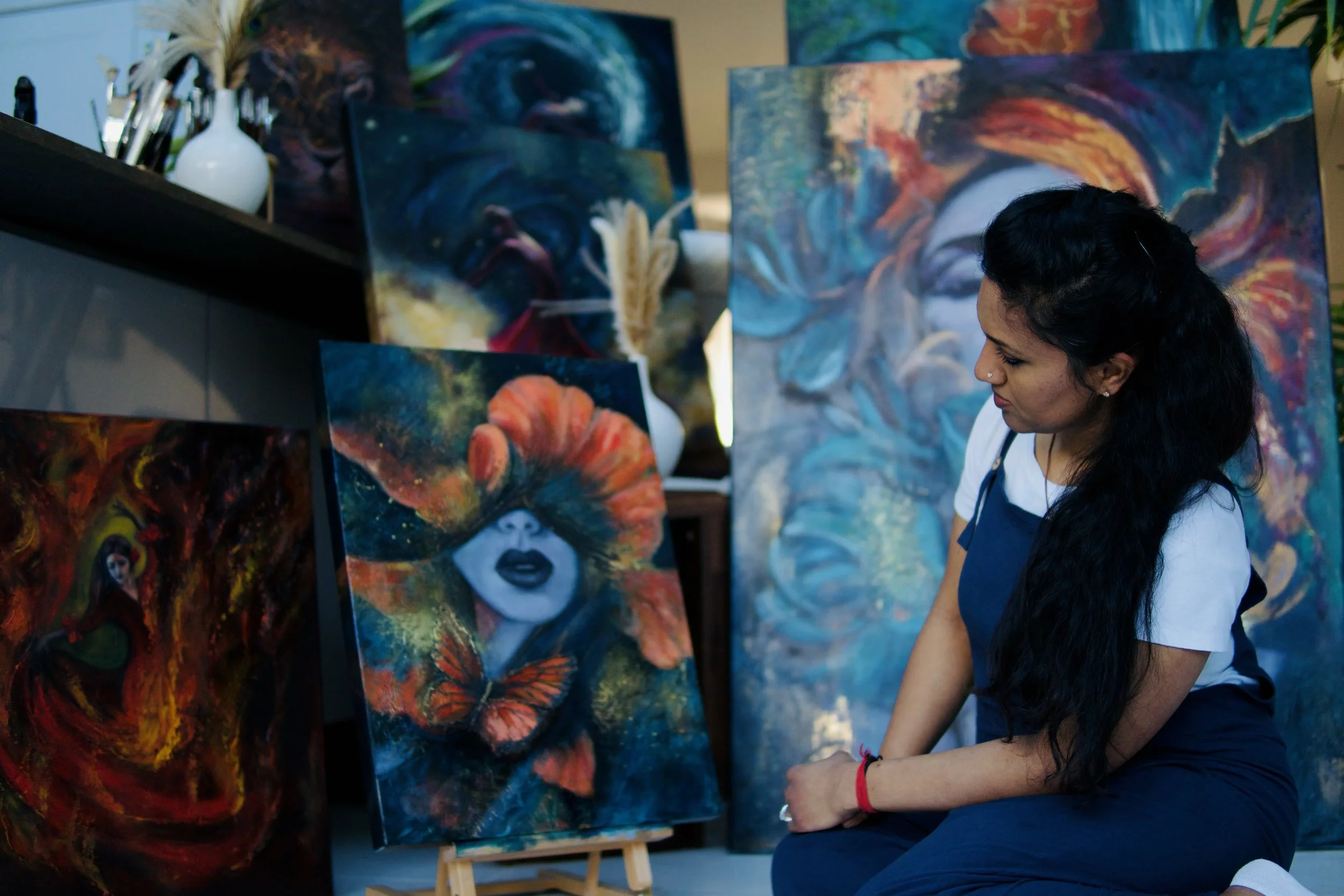 Woman with long dark hair and nose piercing looking at abstract paintings, including a portrait of a woman with a colorful headdress, in an art gallery or studio.