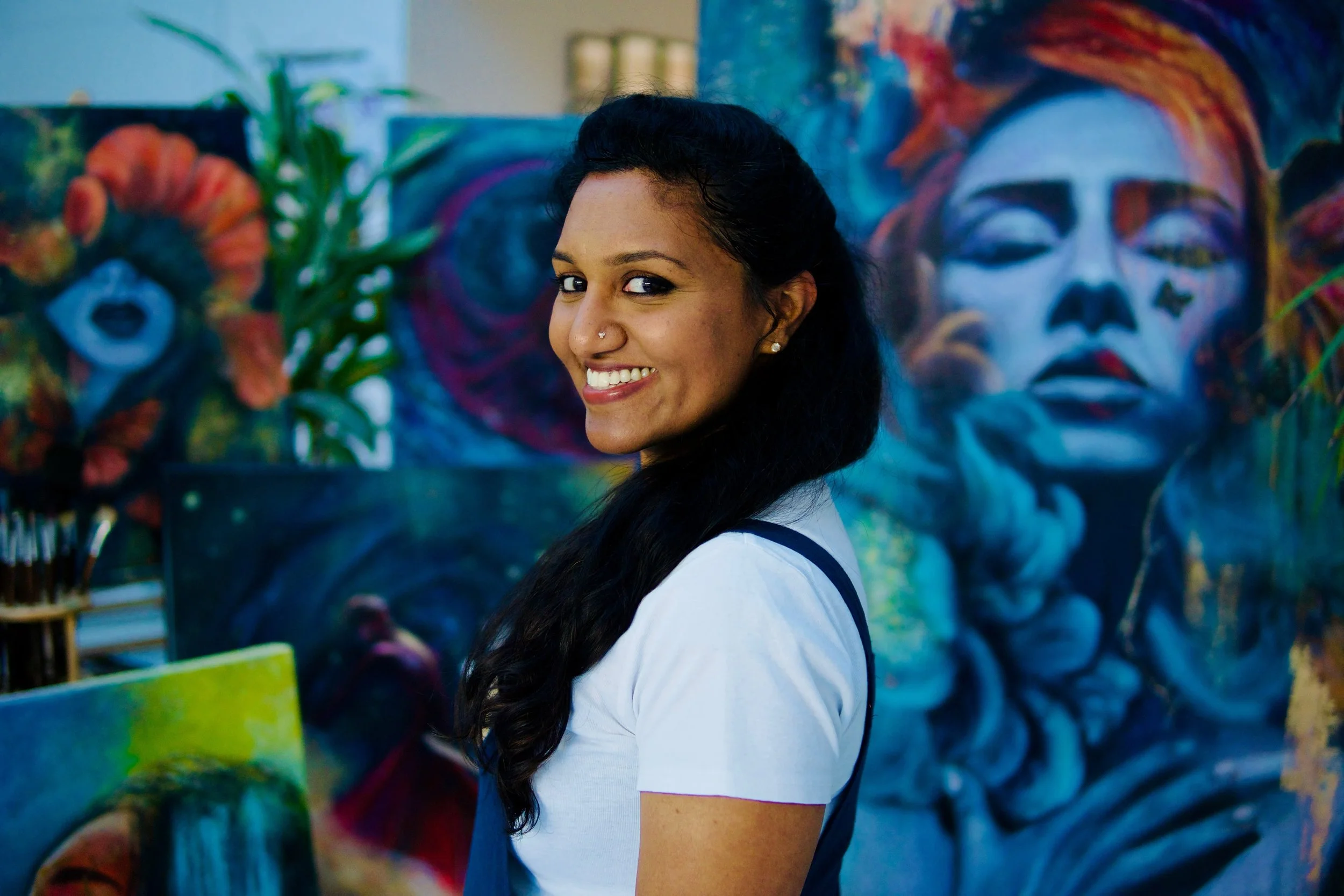 A woman with long dark hair, smiling, standing near colorful paintings of faces and abstract art at an art gallery.