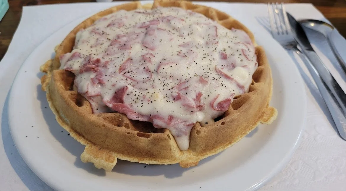 Waffle topped with ham and creamed chipped beef, sprinkled with black pepper, on a white plate with utensils beside it.