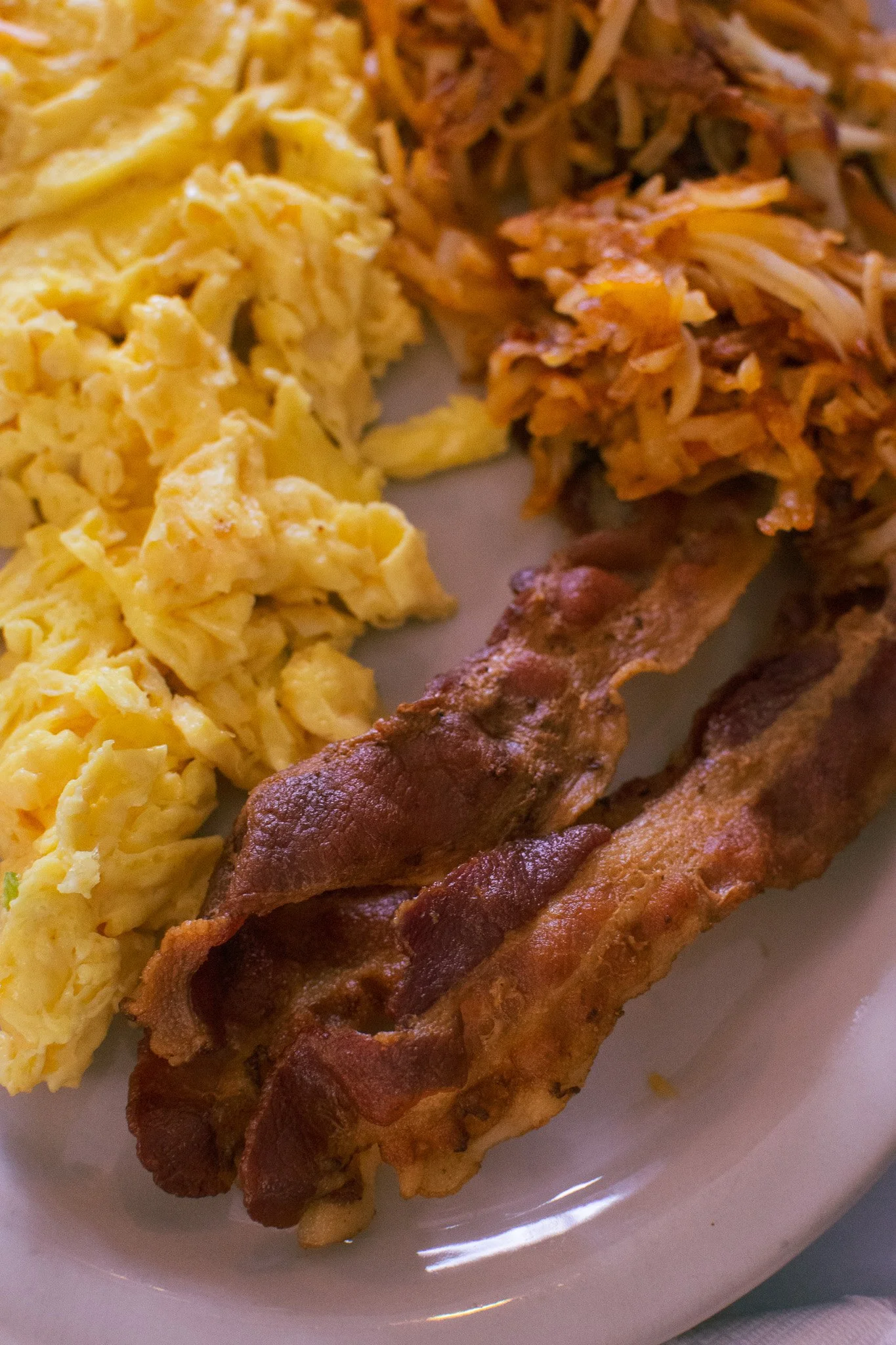 Scrambled eggs, hash browns, and bacon on a white plate.
