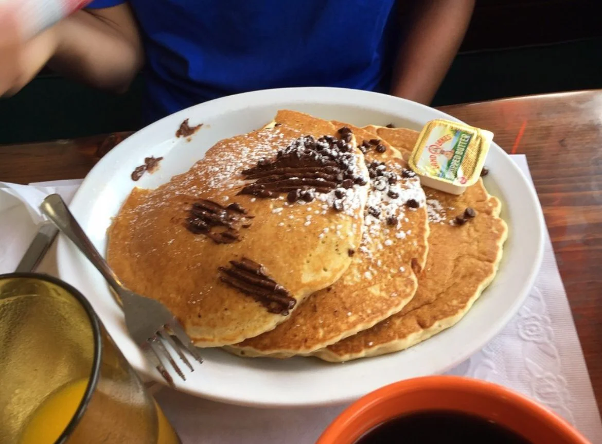 A plate of three large pancakes topped with chocolate chips, powdered sugar, and syrup, with a butter packet on the side, next to a cup of coffee and a glass of orange juice.