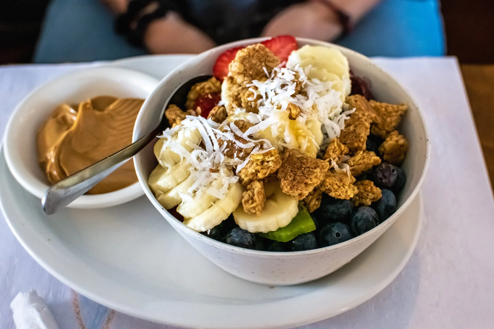 A bowl of acai with granola, sliced bananas, blueberries, shredded coconut, and a serving of peanut butter on the side.
