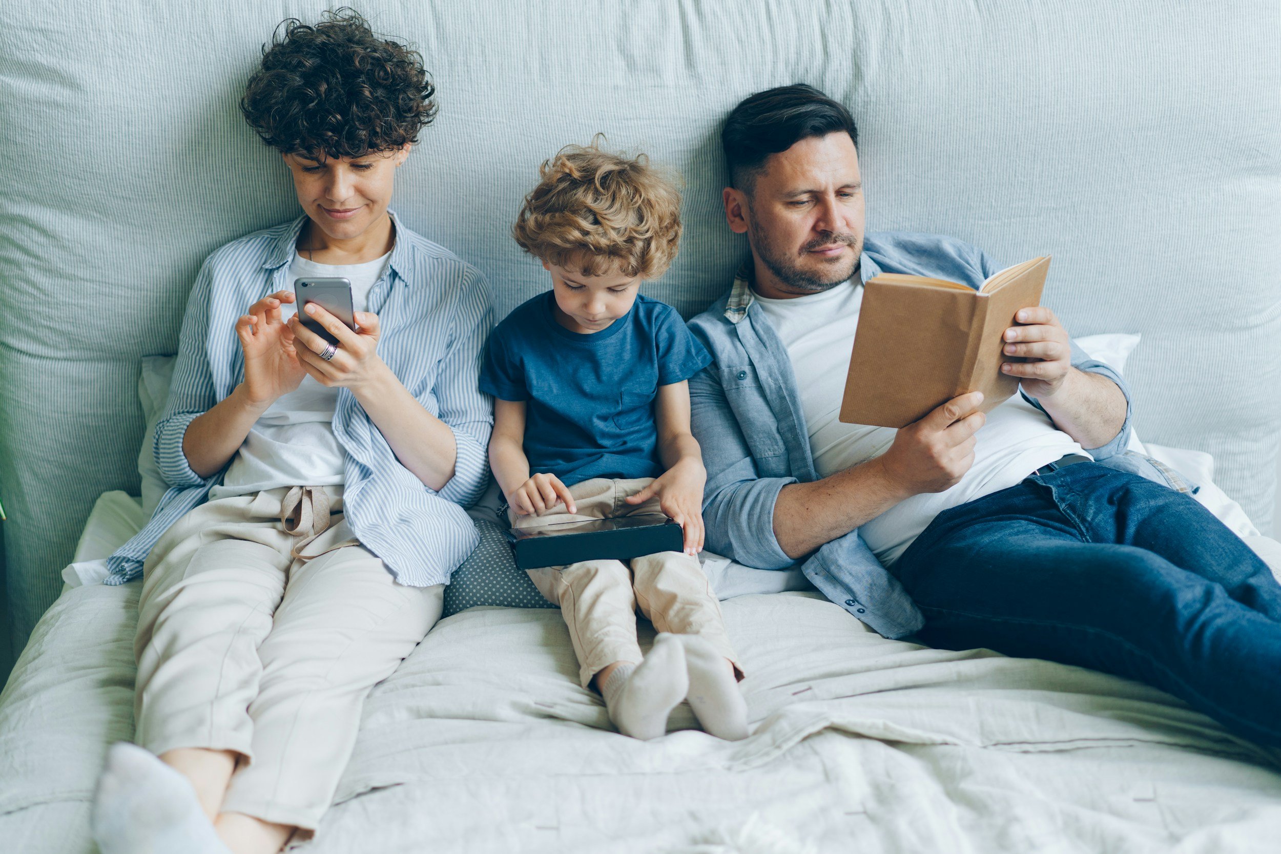A family of three sitting on a bed, with the mother on the left using a smartphone, the young boy in the middle looking down at a tablet, and the father on the right reading a book.