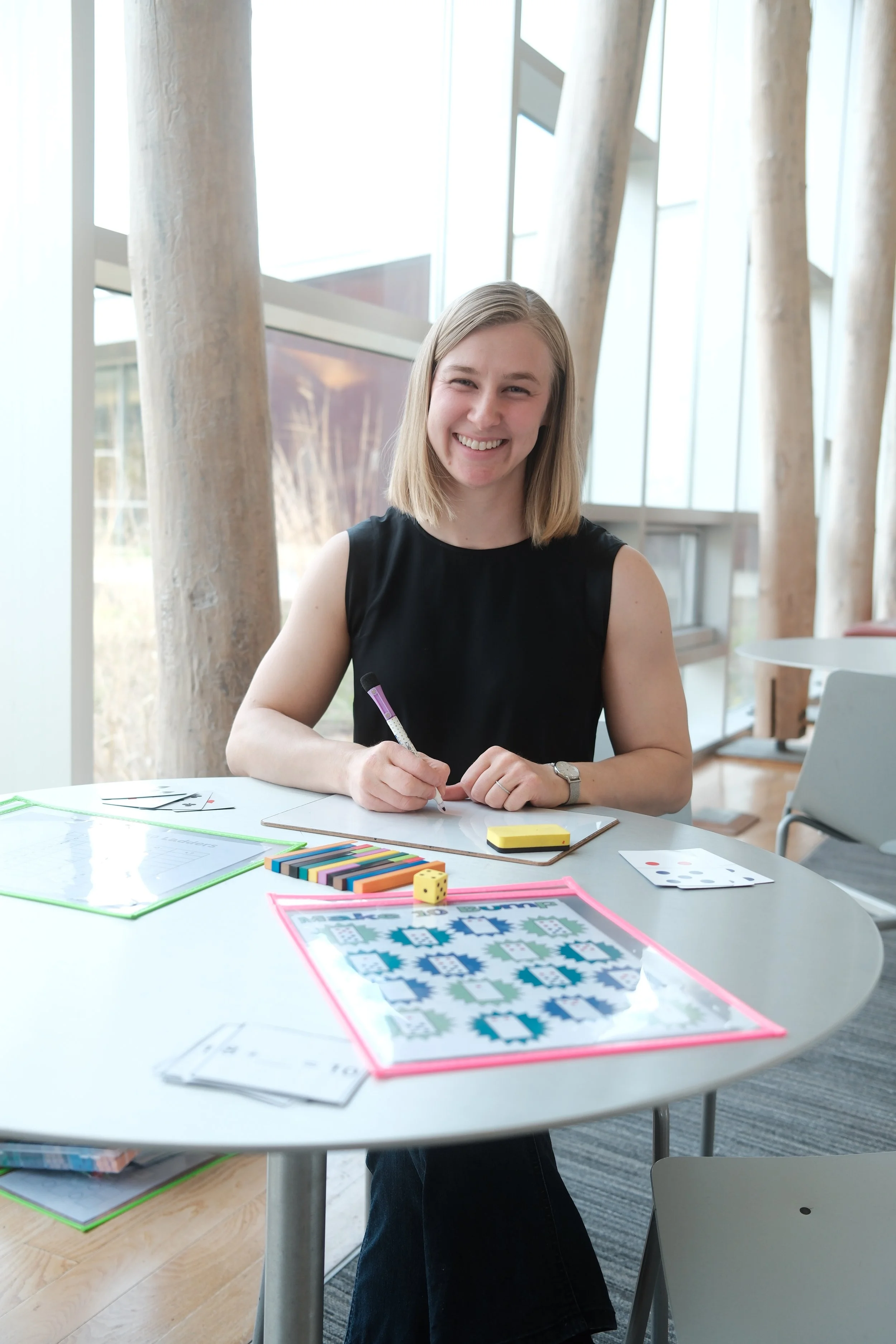 A smiling young woman sitting at a round table with board game components, including game cards, dice, and colored pieces, in a bright modern indoor space with large windows and wooden beams.