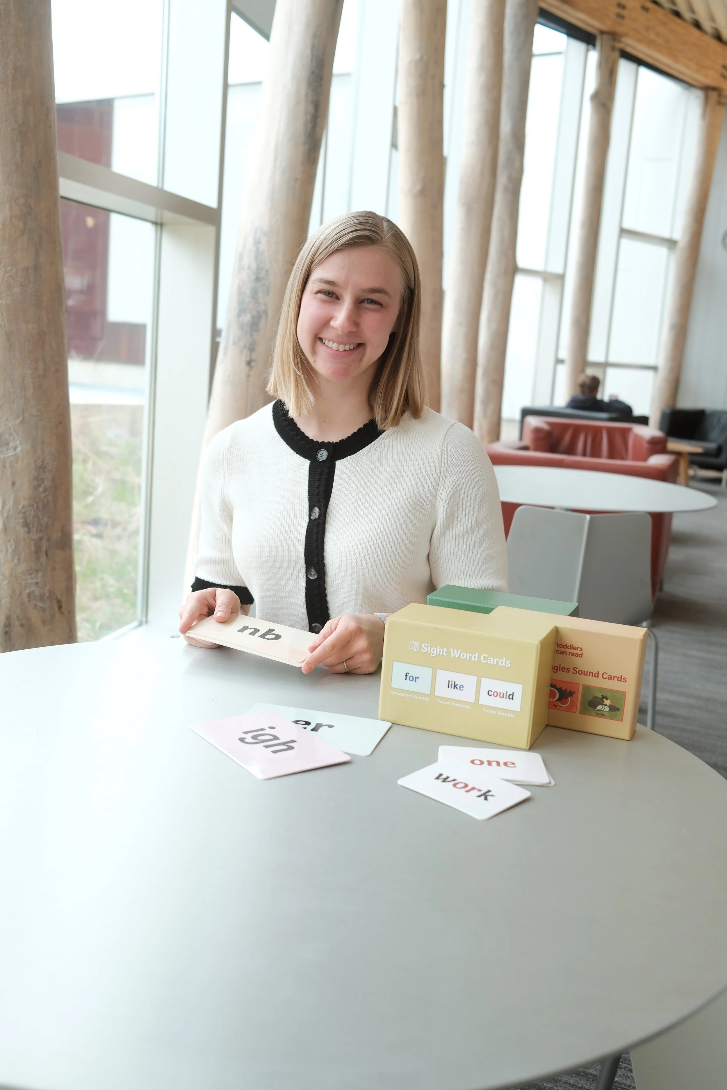 A smiling young woman with shoulder-length blonde hair sitting at a table, holding a flash card with the letters 'nb' on it. There are sight word cards on the table with words like 'high,' 'one,' 'work,' and others, and educational boxes labeled 'Sight Word Cards' and 'tiggies Sound Cards.' The background features large windows, wooden beams, and cozy seating areas.