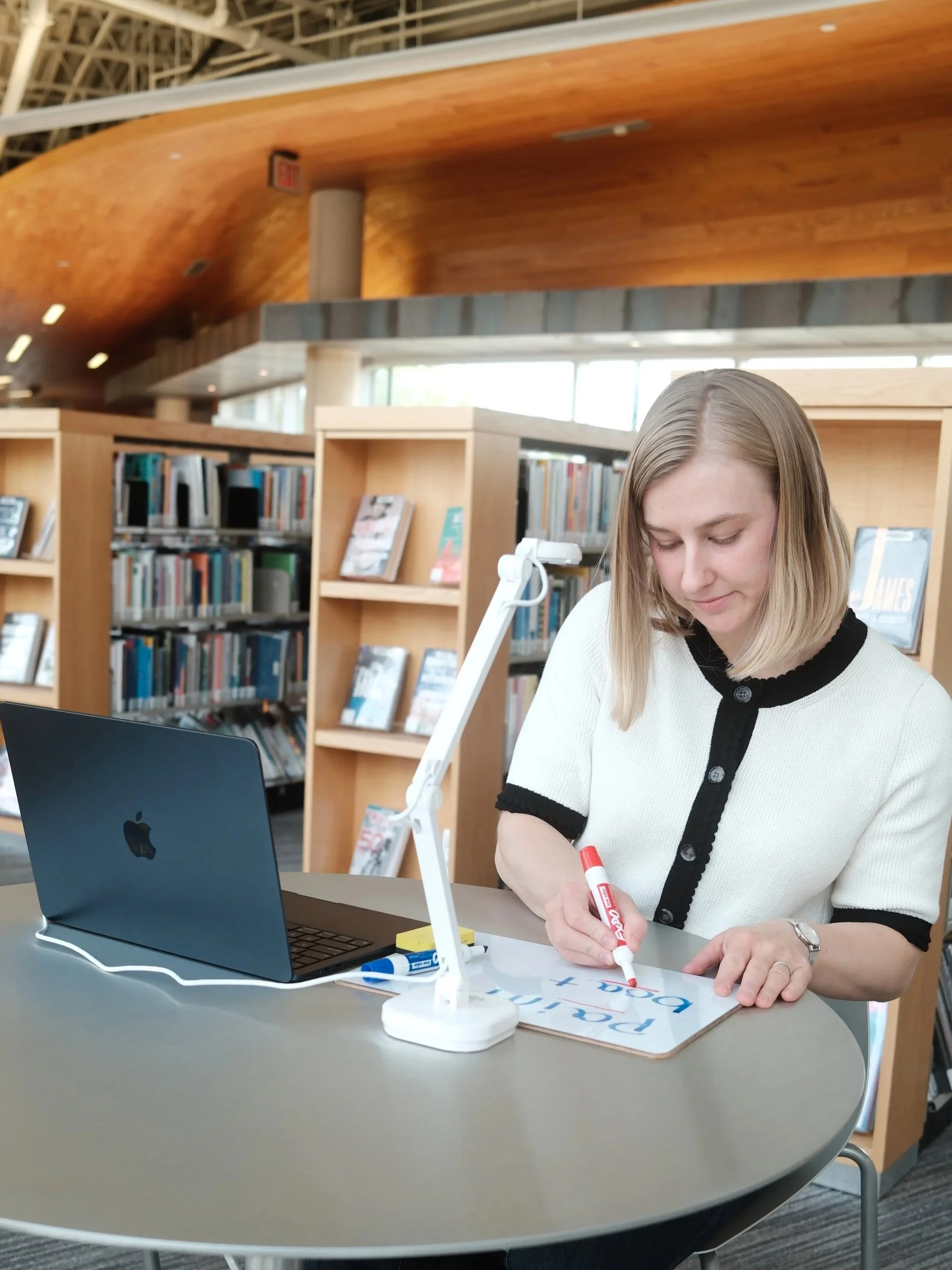 A woman sitting at a table in a library, writing with a red marker on a whiteboard or poster, with a silver MacBook laptop and a white desk lamp on the table, and bookshelves filled with books in the background.