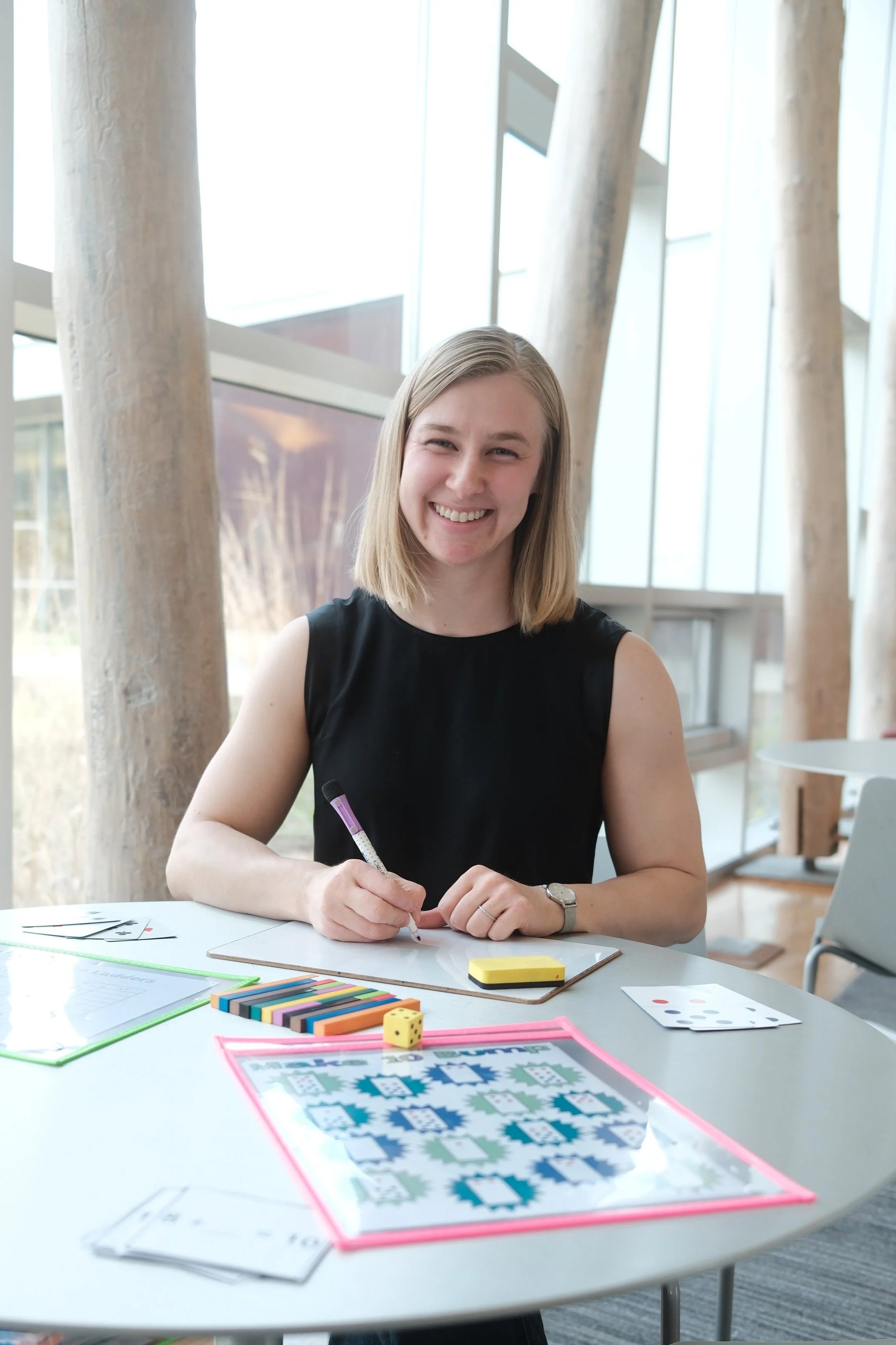Young woman with blonde hair smiling and sitting at a table with game pieces, dice, and playing cards, in a well-lit modern building with large windows and curtains.