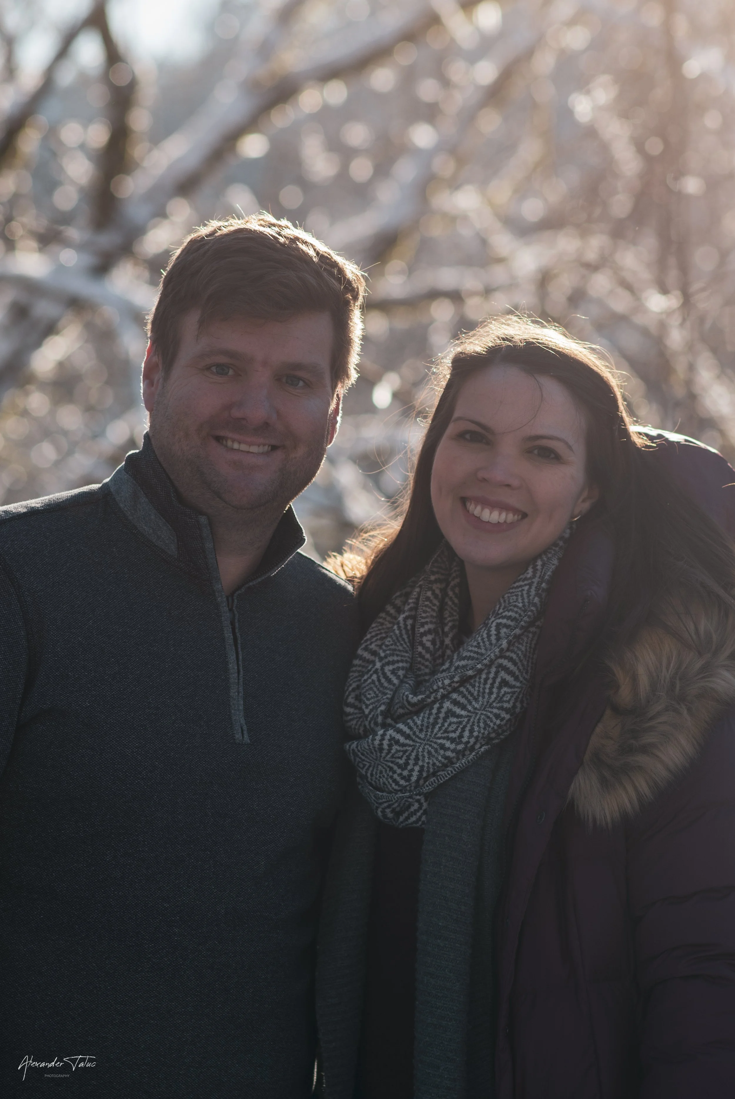 A smiling man and woman outdoors in winter, with sunlight and snow-covered trees in the background.